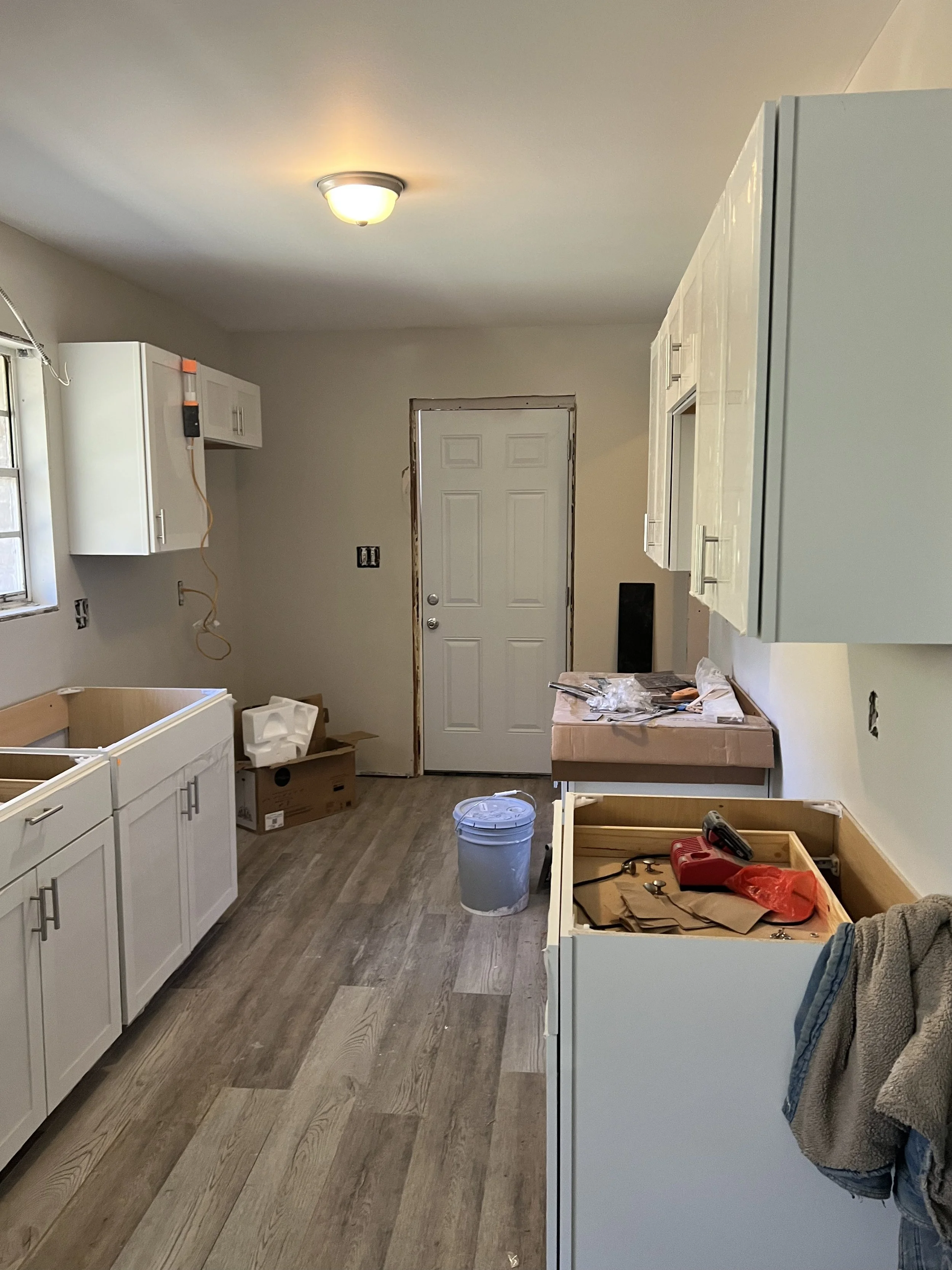 Kitchen under renovation with unfinished cabinets, tools, and construction materials scattered around, a ceiling light, and a door at the back.