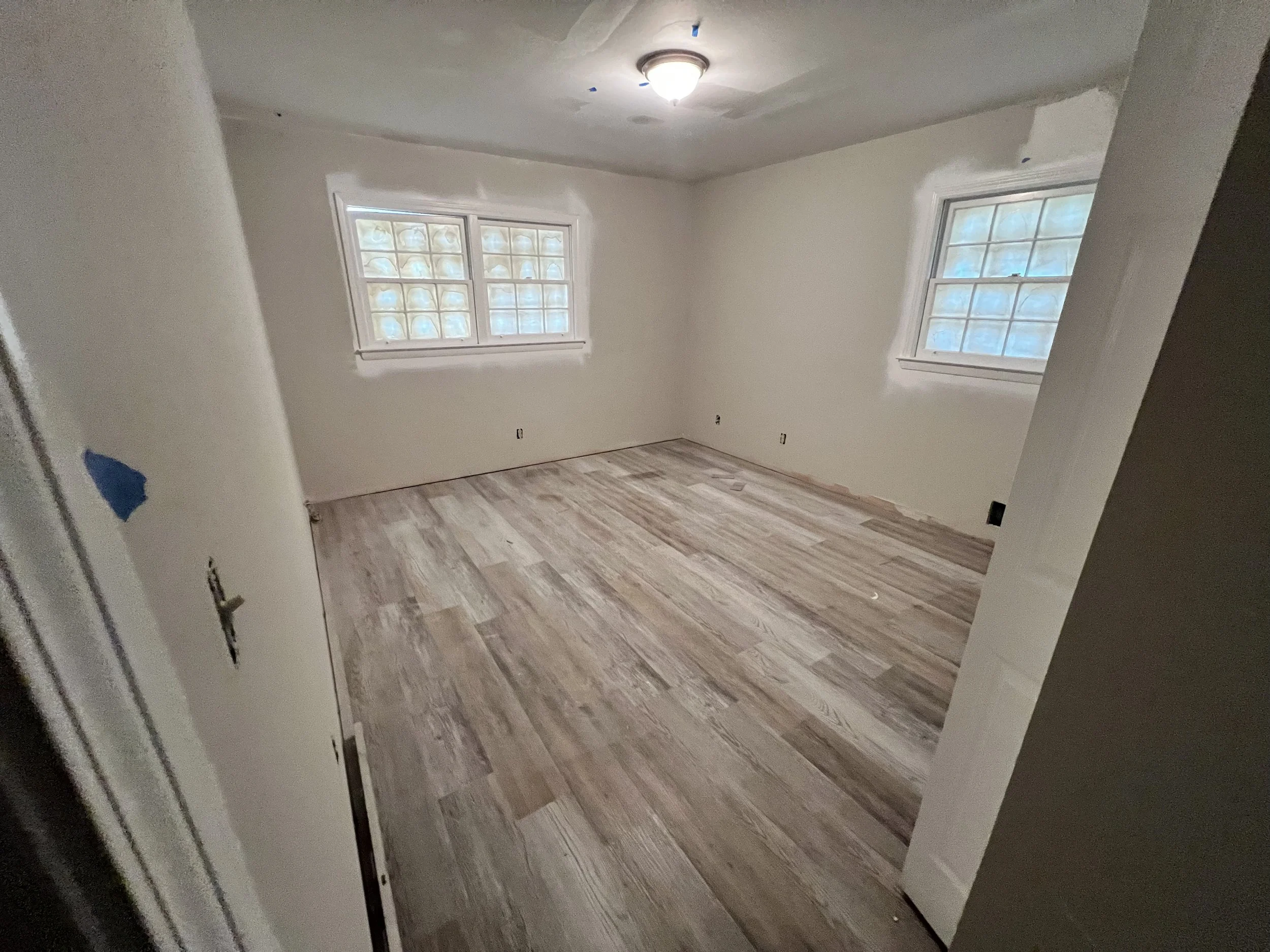 An empty room with light-colored walls, hardwood flooring, two windows with frosted glass blocks, and a ceiling light fixture.