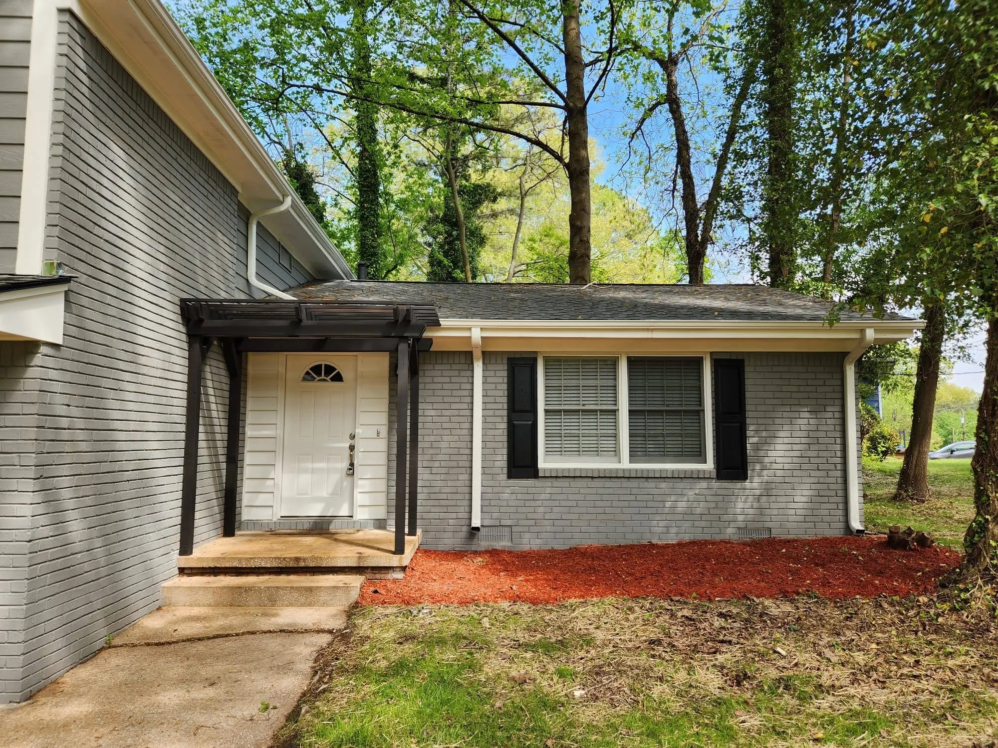 Front view of a house with a white door, black shutters, gray brick exterior, and a small porch with steps, surrounded by trees.