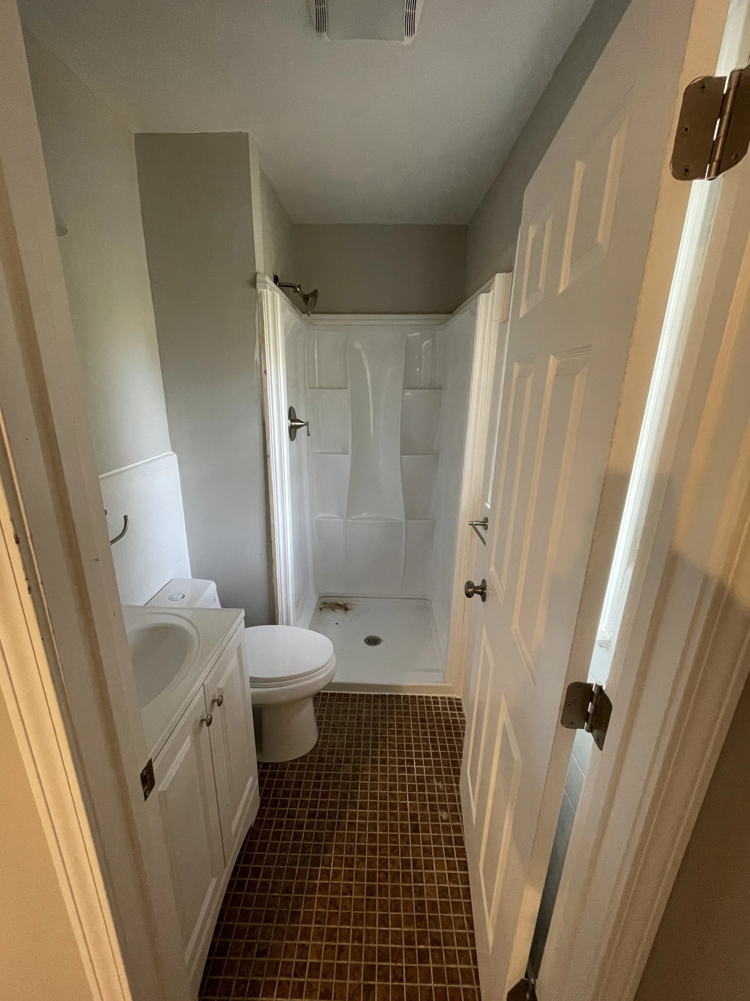 Small bathroom with a shower, toilet, and a small sink with a cabinet. Brown tile floor and gray walls.