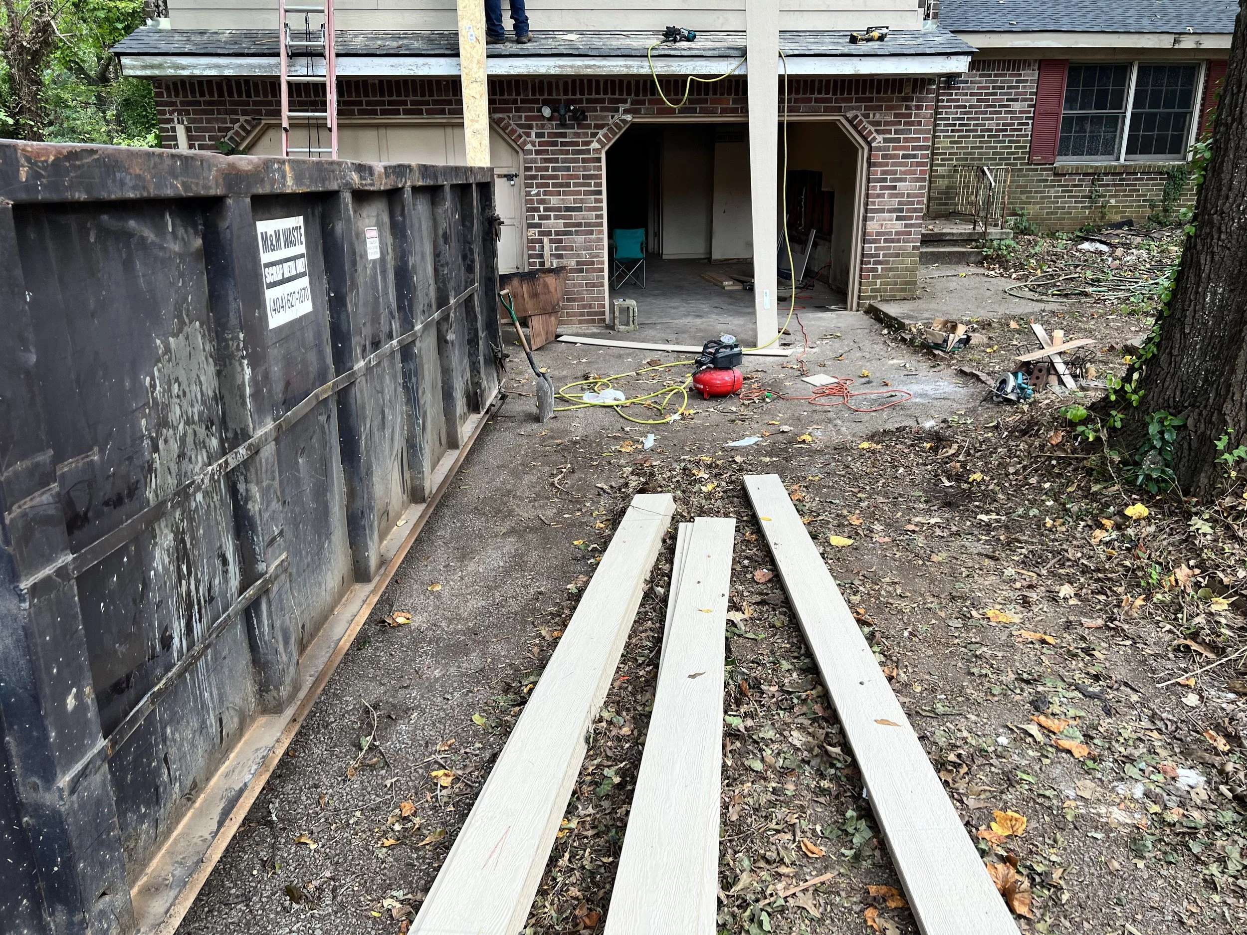 Construction site in the backyard of a house with building materials, tools, and debris, partially built porch, and trees around.