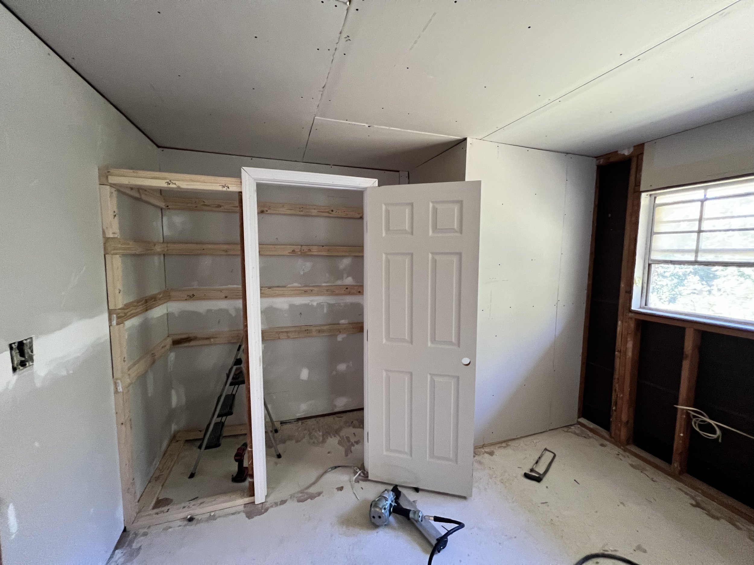 Room under construction with unfinished walls, an open closet with wooden framing, a partially installed door, a window to the right, and construction tools on the floor.