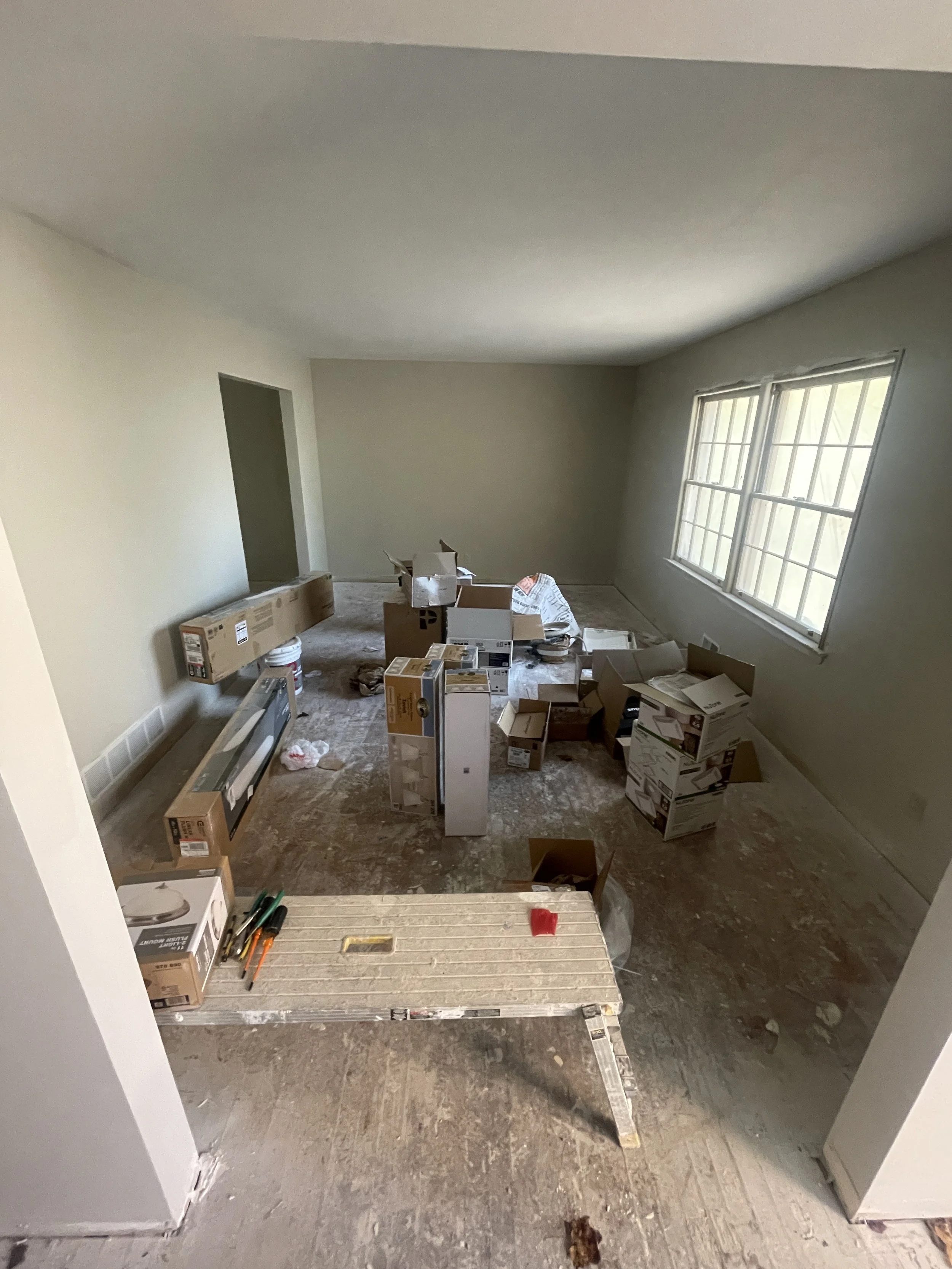 Empty room under renovation with cardboard boxes, tools, and construction materials scattered on the floor, large window on the right side, and unfinished floor and walls.