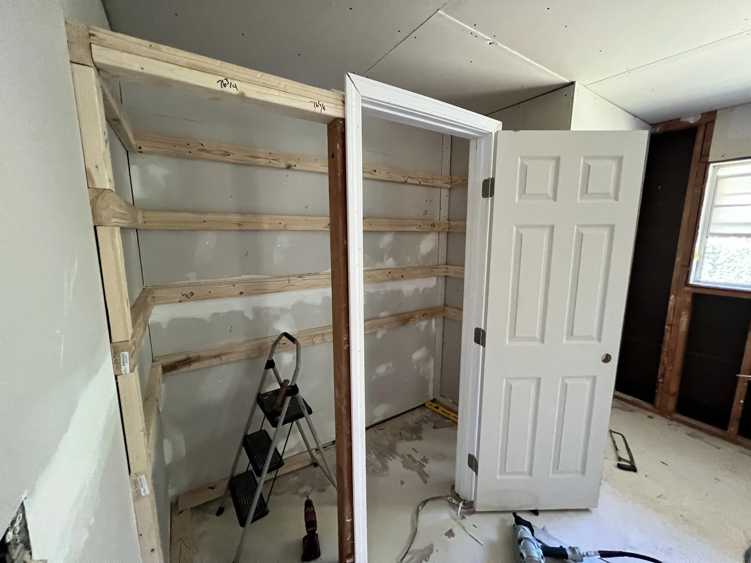 Interior of a room under construction showing a partially built wooden closet frame, an open white door, unfinished walls, construction tools and materials on the floor, and a window with daylight coming through.