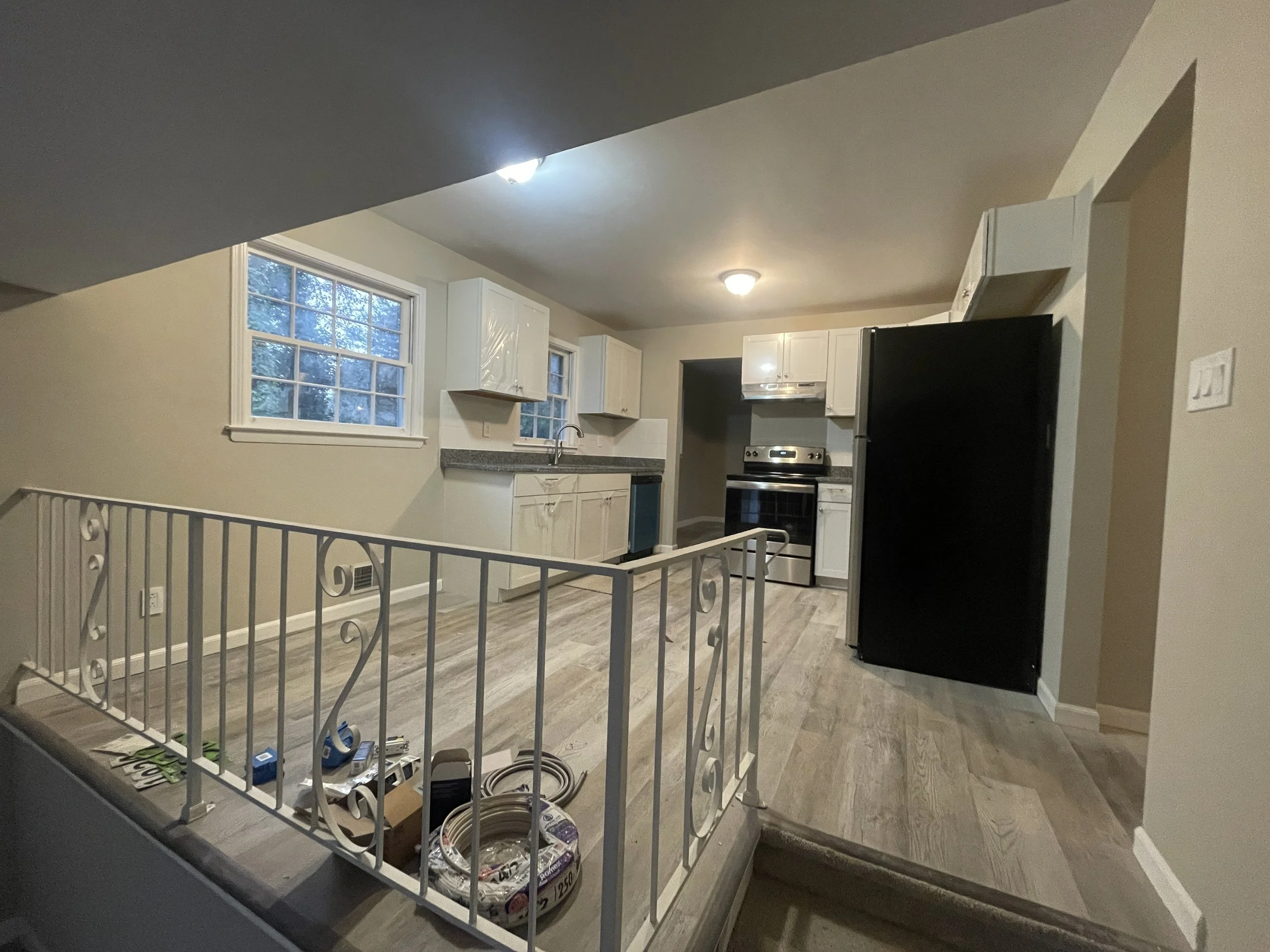 Empty kitchen with white cabinets, black refrigerator, and stainless steel stove, with wood flooring and two windows, taken from a slightly elevated perspective.