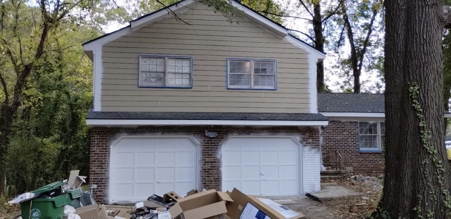 A two-story house with a failing in the garage area. The exterior has beige siding on the second floor and brick on the first floor. There are two garage doors and several windows. Debris and garbage bins are scattered in front of the house, suggesti
