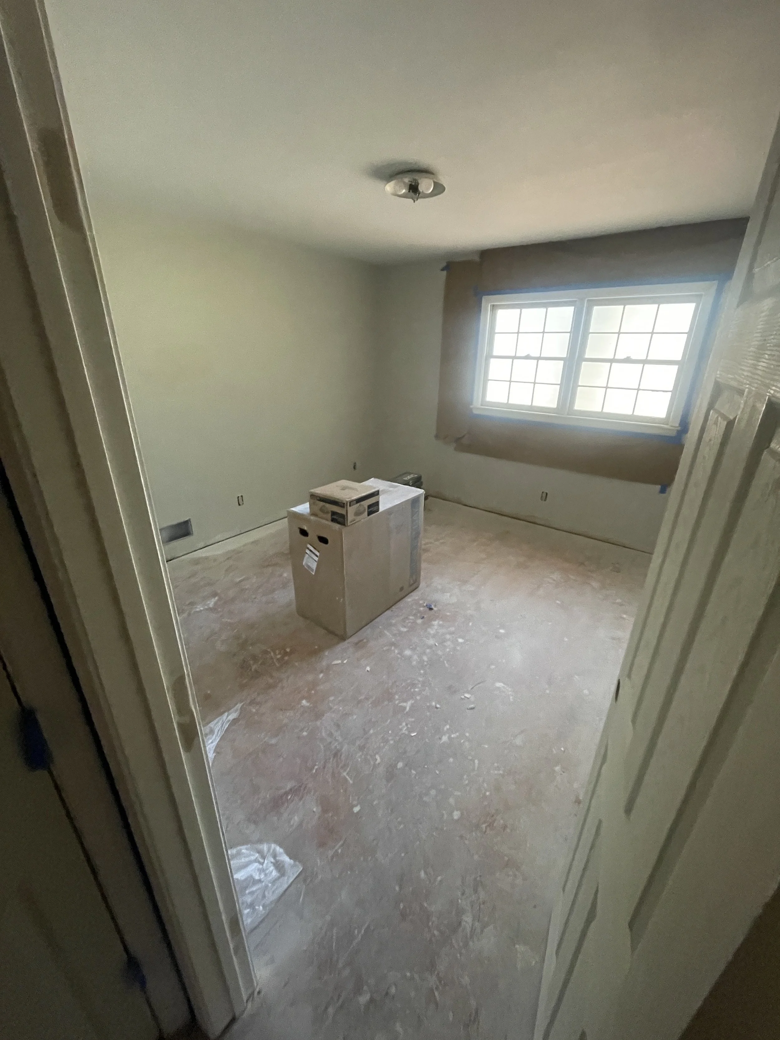 Empty room under renovation with a window covered in brown paper, a cardboard box on the unfinished floor, and marks on the walls.