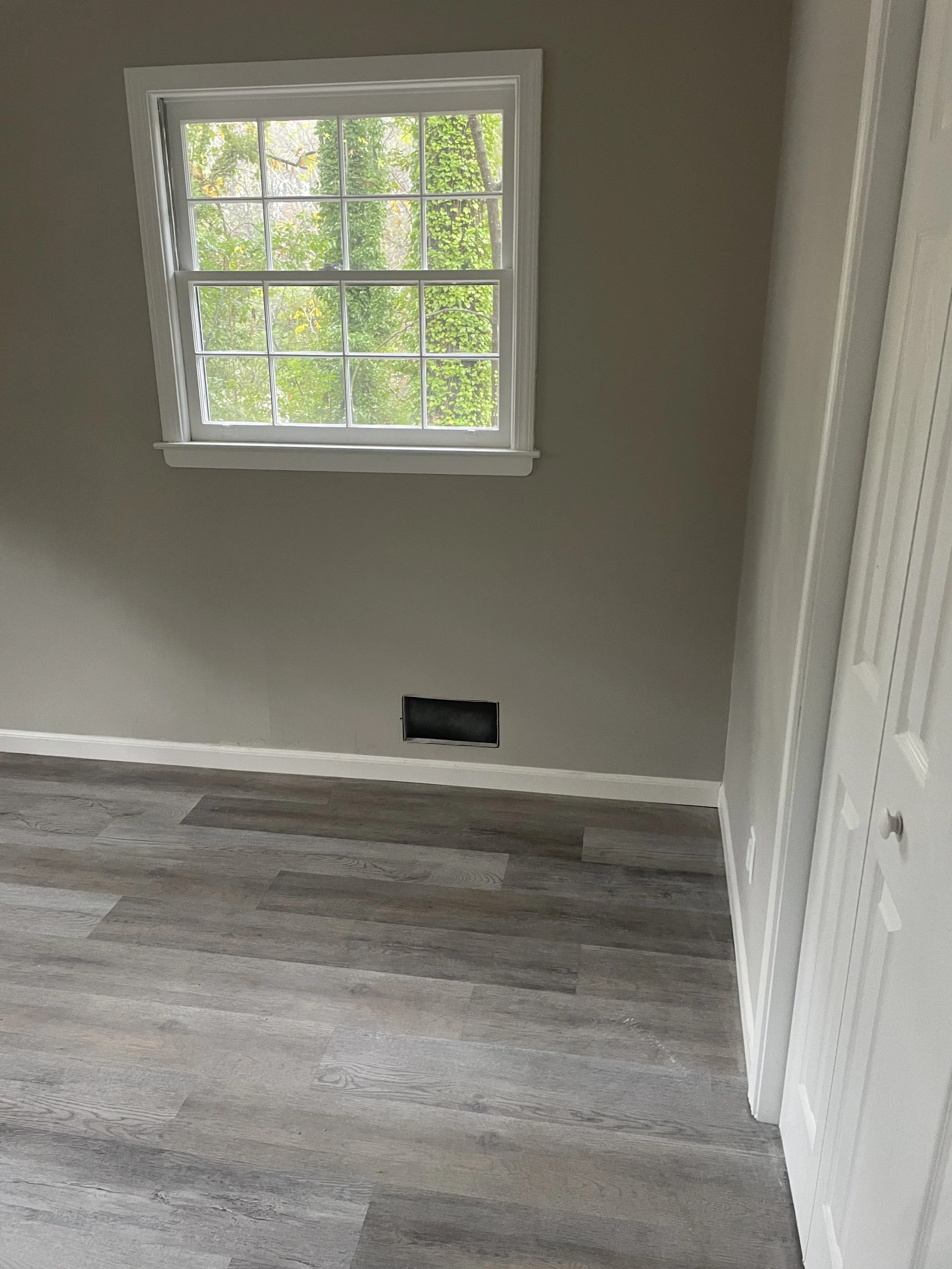 An empty room with light gray walls, a window with multiple panes showing green trees outside, a black vent, and white trim around the window and baseboards.