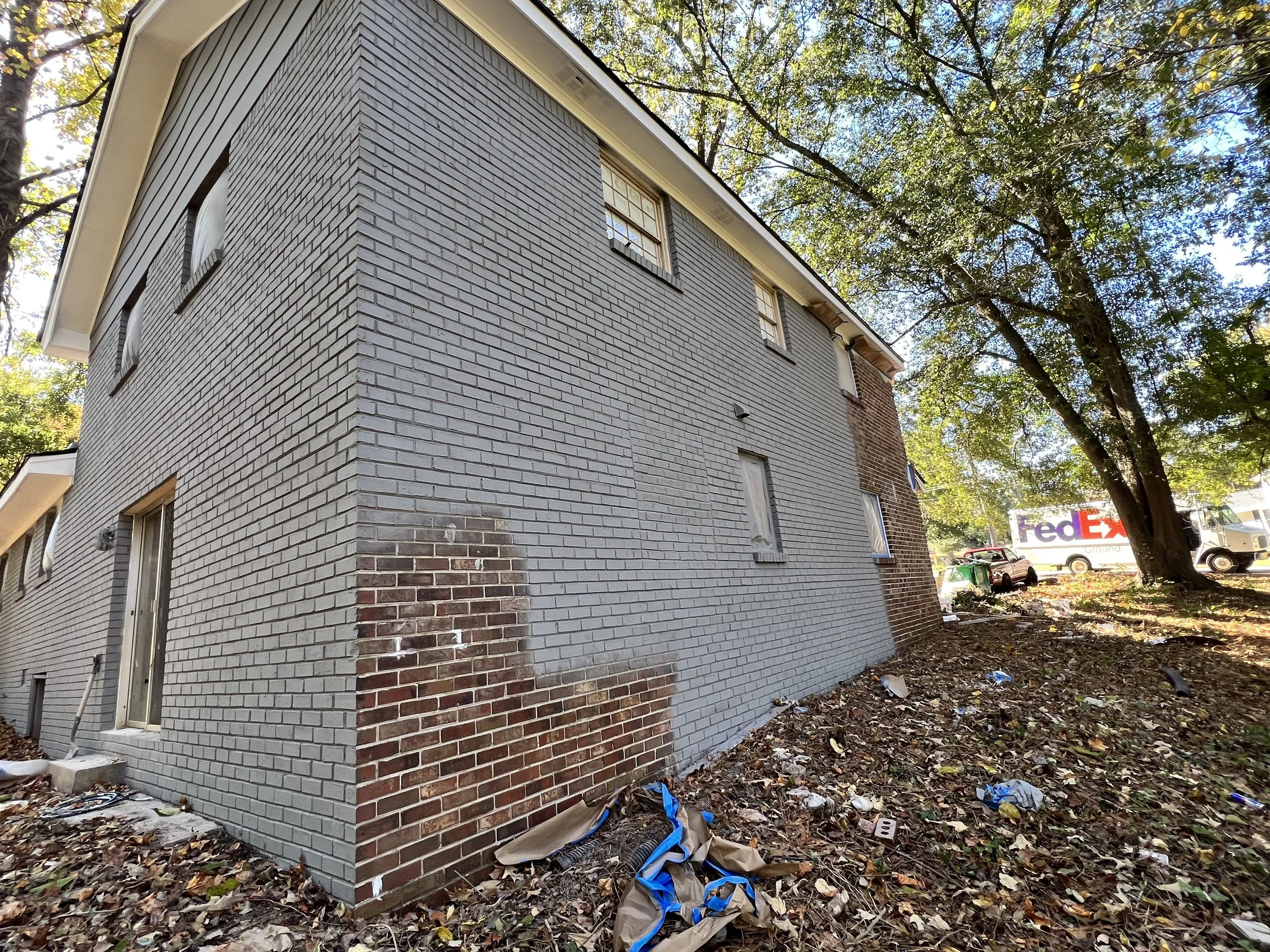 Side view of a gray brick building with a new section, some windows covered with plastic, surrounded by leaf-covered ground and a large leafy tree, with parked trucks and a FedEx delivery truck in the background.