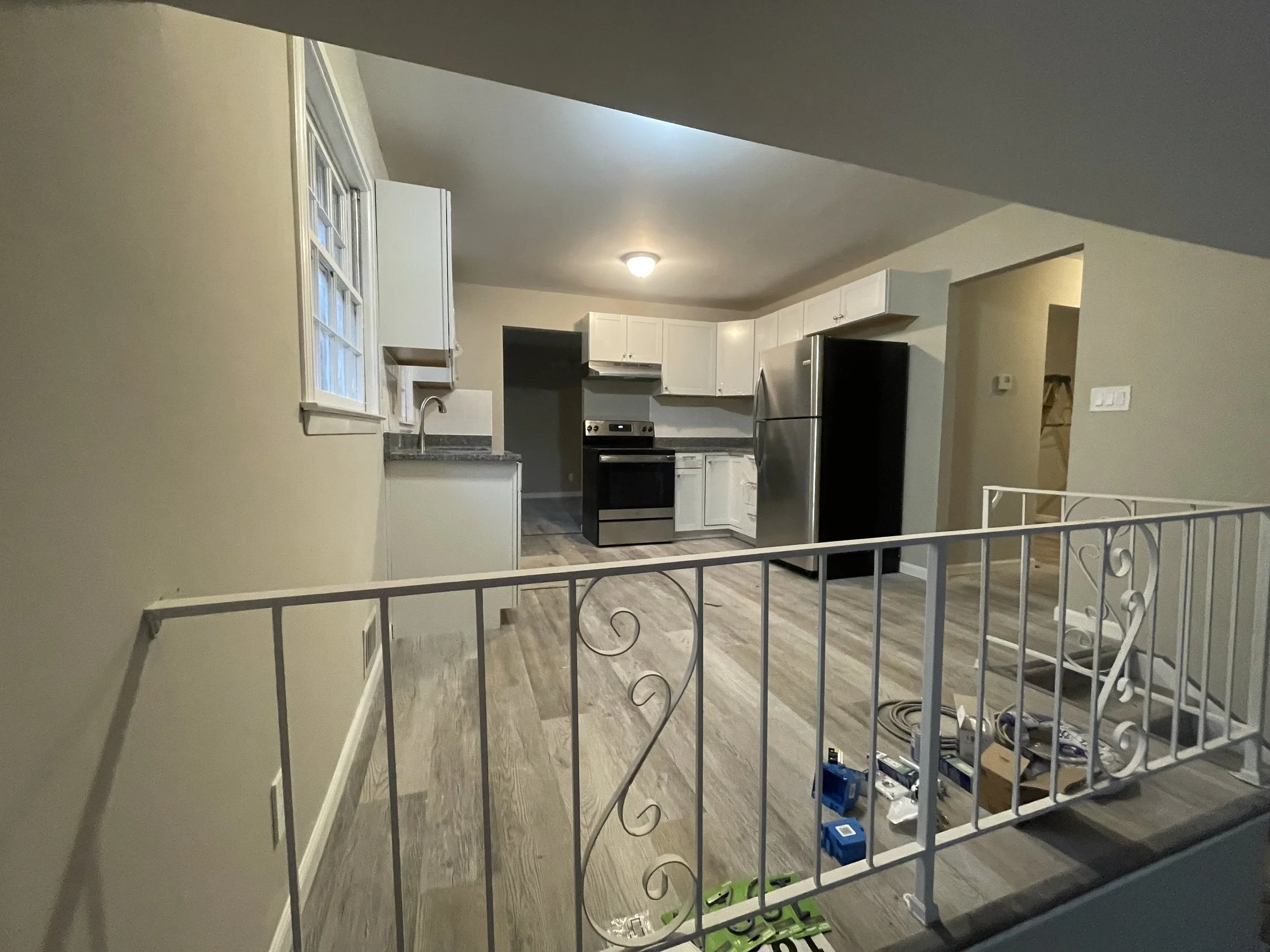 Empty kitchen with white cabinets, stainless steel refrigerator, oven, and double sink. Light-colored wood flooring, beige walls, with tools and materials on the floor, and a white wrought iron staircase railing in the foreground.