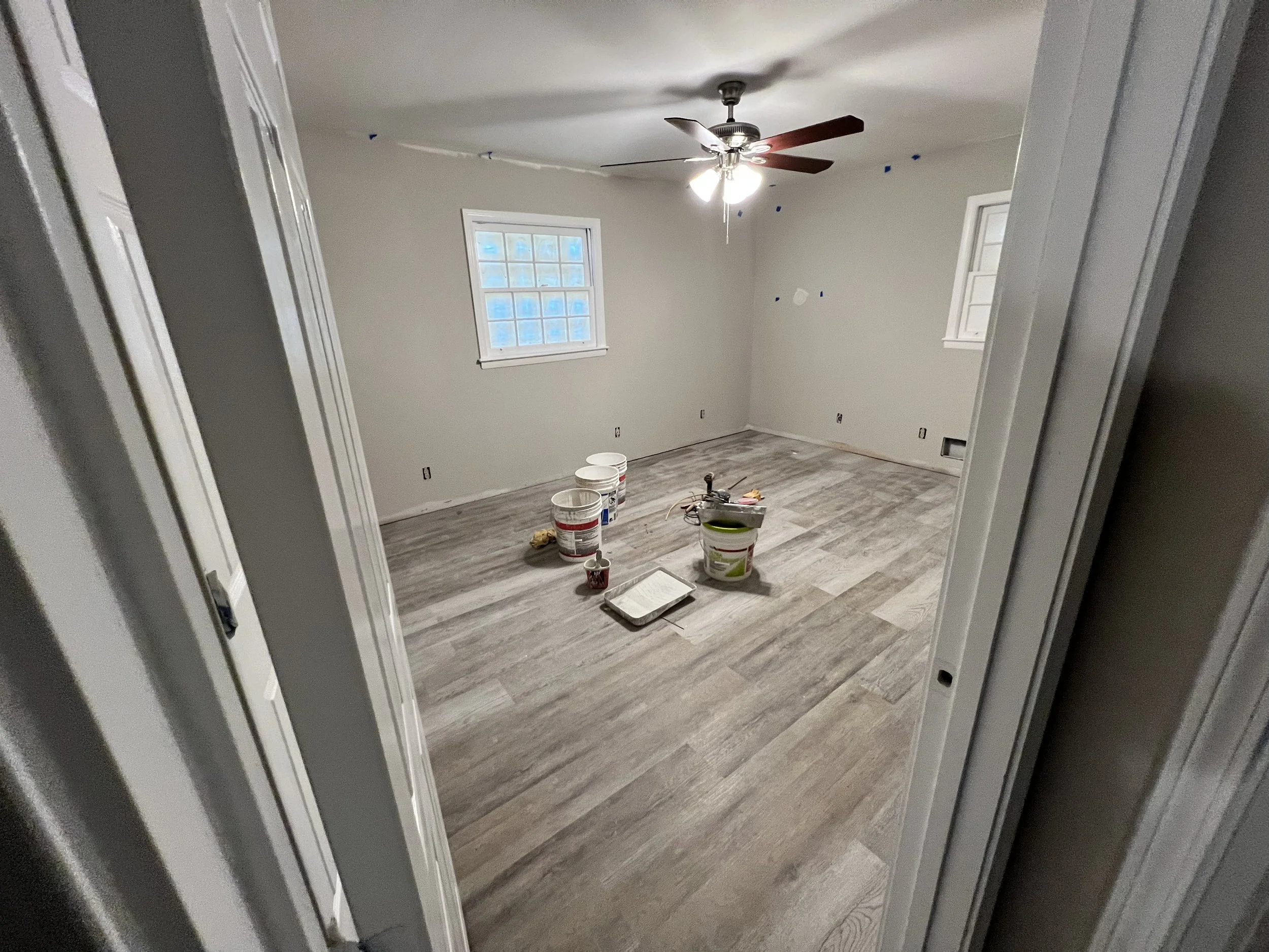 An empty room under renovation with light-colored wooden flooring, two windows, and a ceiling fan with lights. There are buckets, a paint roller tray, and other tools on the floor.