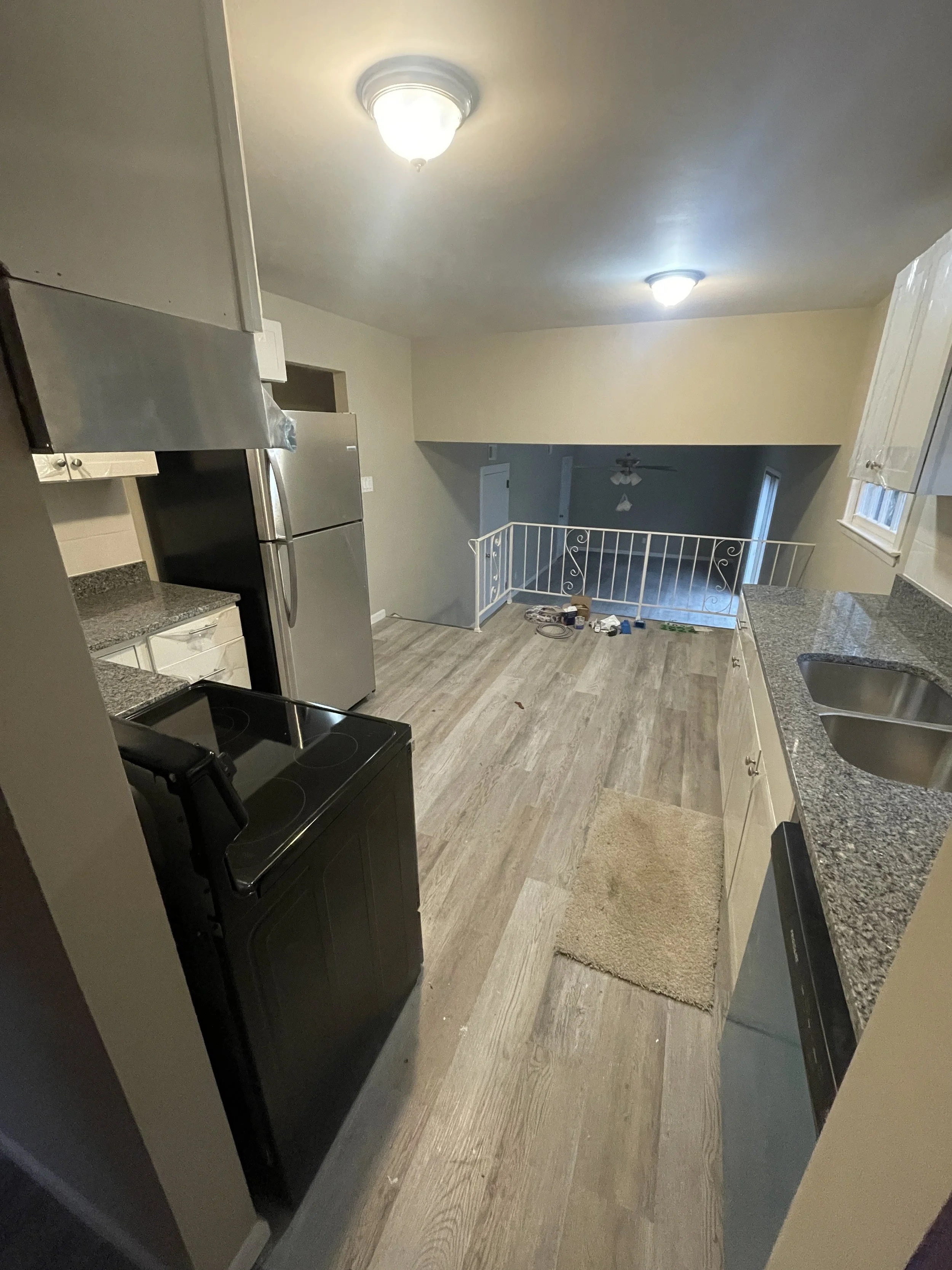 Empty kitchen with granite countertops, white cabinets, a stainless steel refrigerator, black stove, sink, and dishwasher. There is a small beige rug on the wooden floor, and some items are near a white railing at the edge of the room.