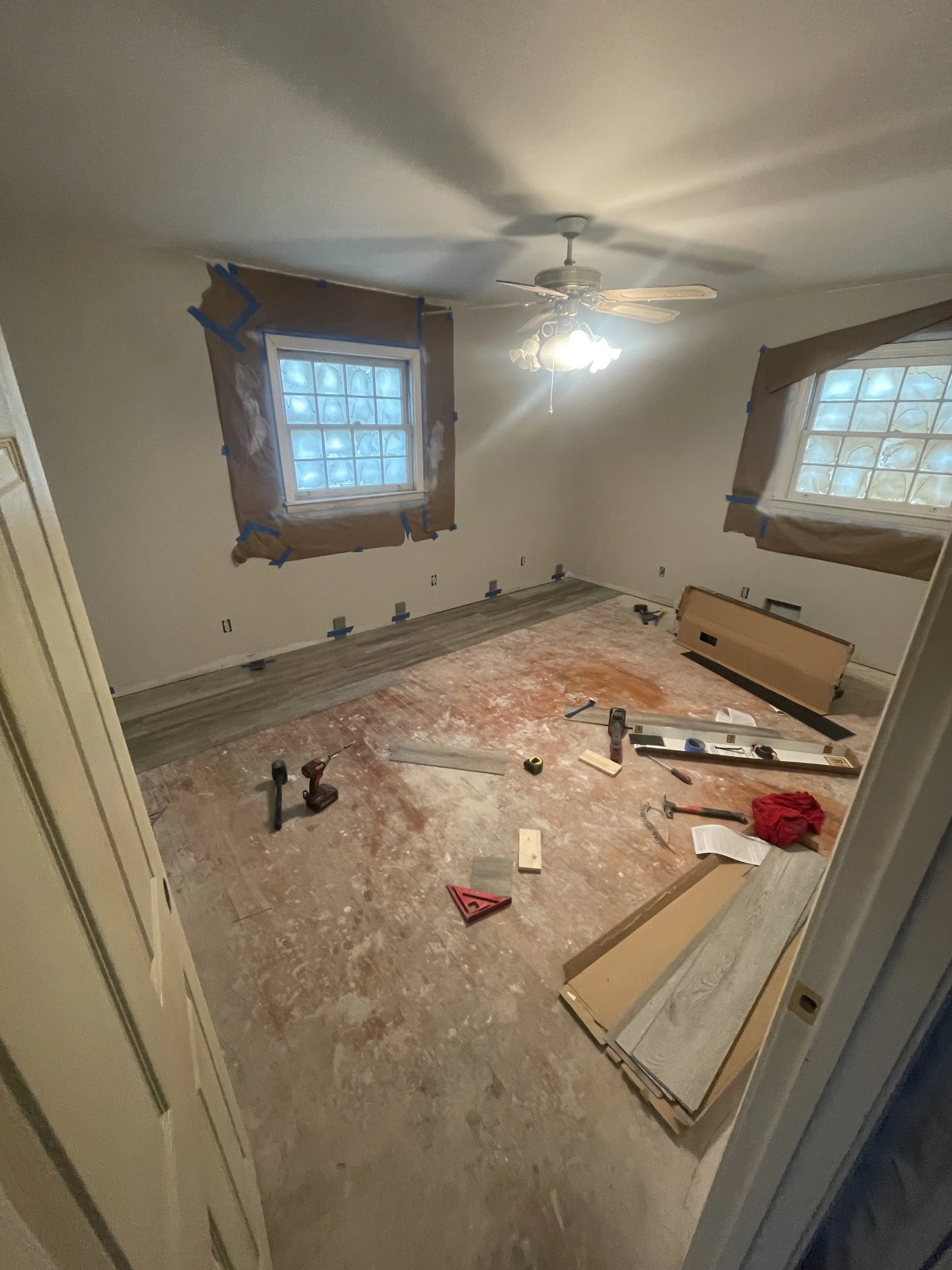 A room under renovation with windows covered in paper and tape, construction tools on the floor, and flooring partially installed.