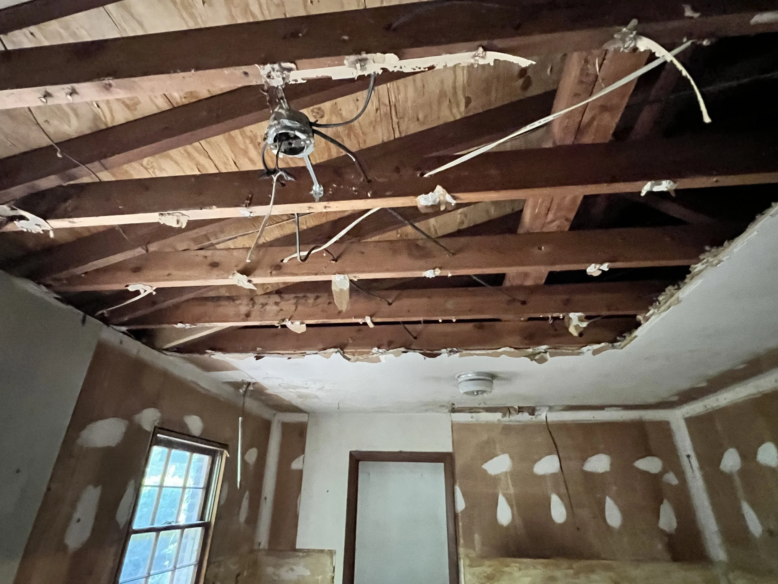 Room with exposed wooden ceiling beams and damaged drywall, with electrical wiring hanging down and a window on the left side.