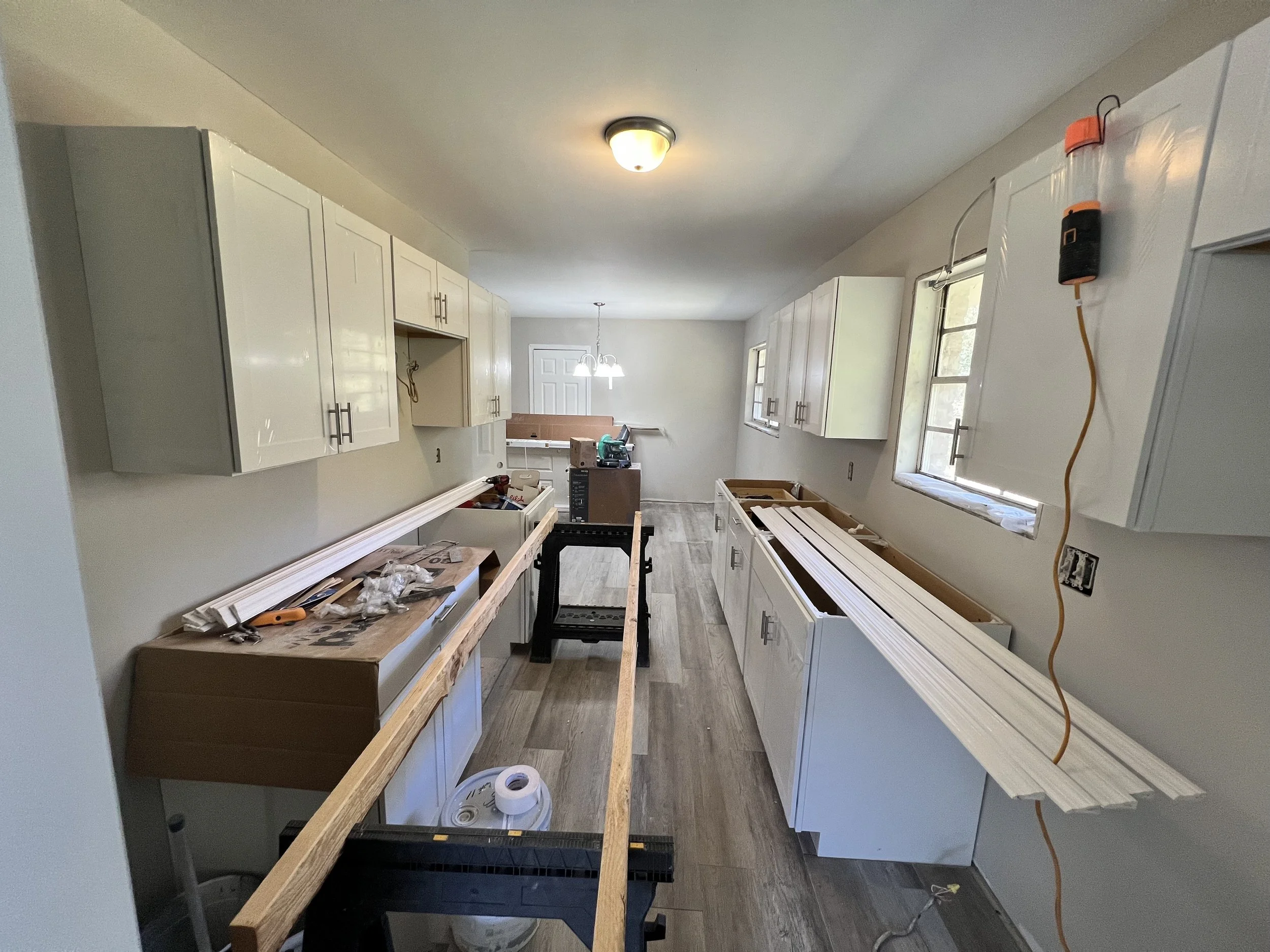 Kitchen under renovation with white upper cabinets on both sides, unfinished lower cabinets and countertop, tools and materials on the counters, and a window on the right wall providing natural light.