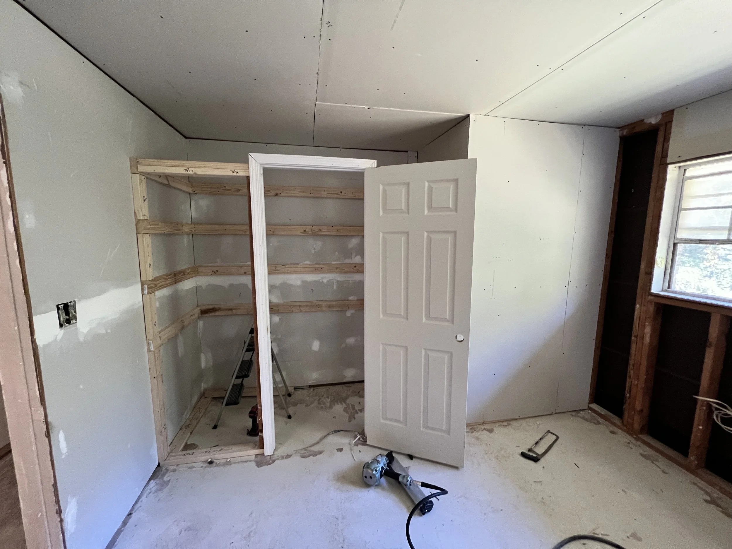 Unfinished room with a newly built wooden shelving unit, a partially open white door, a window on the right side, and construction tools on the floor.