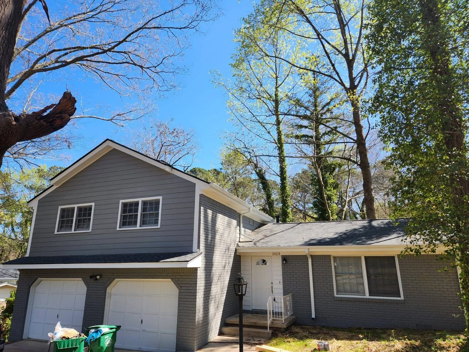 A two-story gray house with white trim, a front porch, and a two-car garage. Trees with early spring foliage surround the house, with leaves just beginning to grow. Two green trash bins are near the garage. The sky is clear and blue.