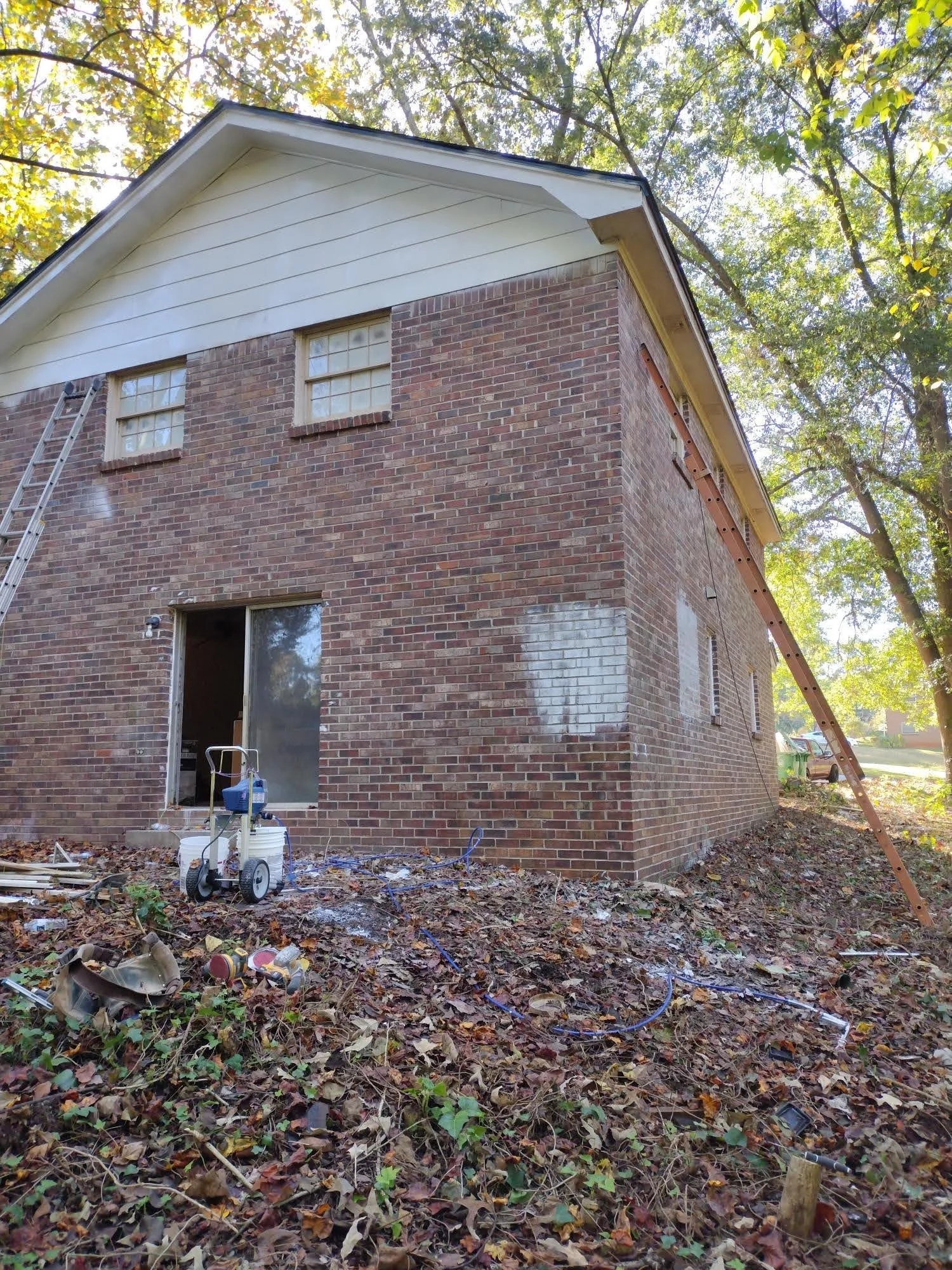 Side view of a brick house under renovation with ladder and construction tools outside, surrounded by fallen leaves.