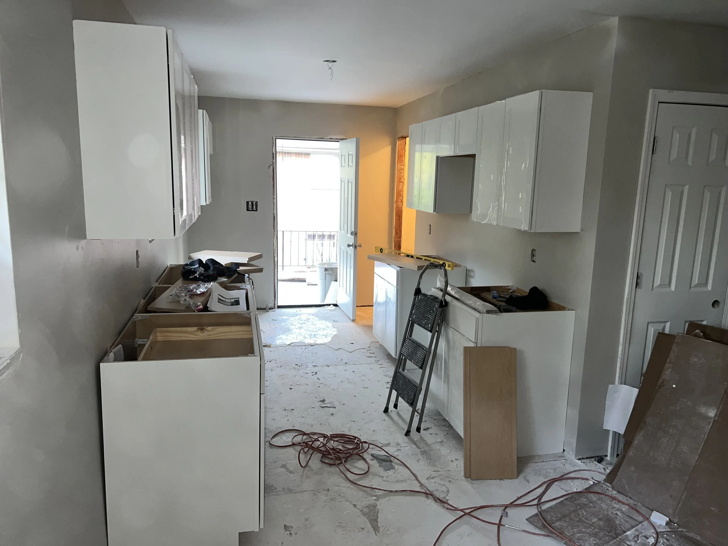 Kitchen under renovation with white cabinets, construction materials, tools, and exposed wiring on the floor.