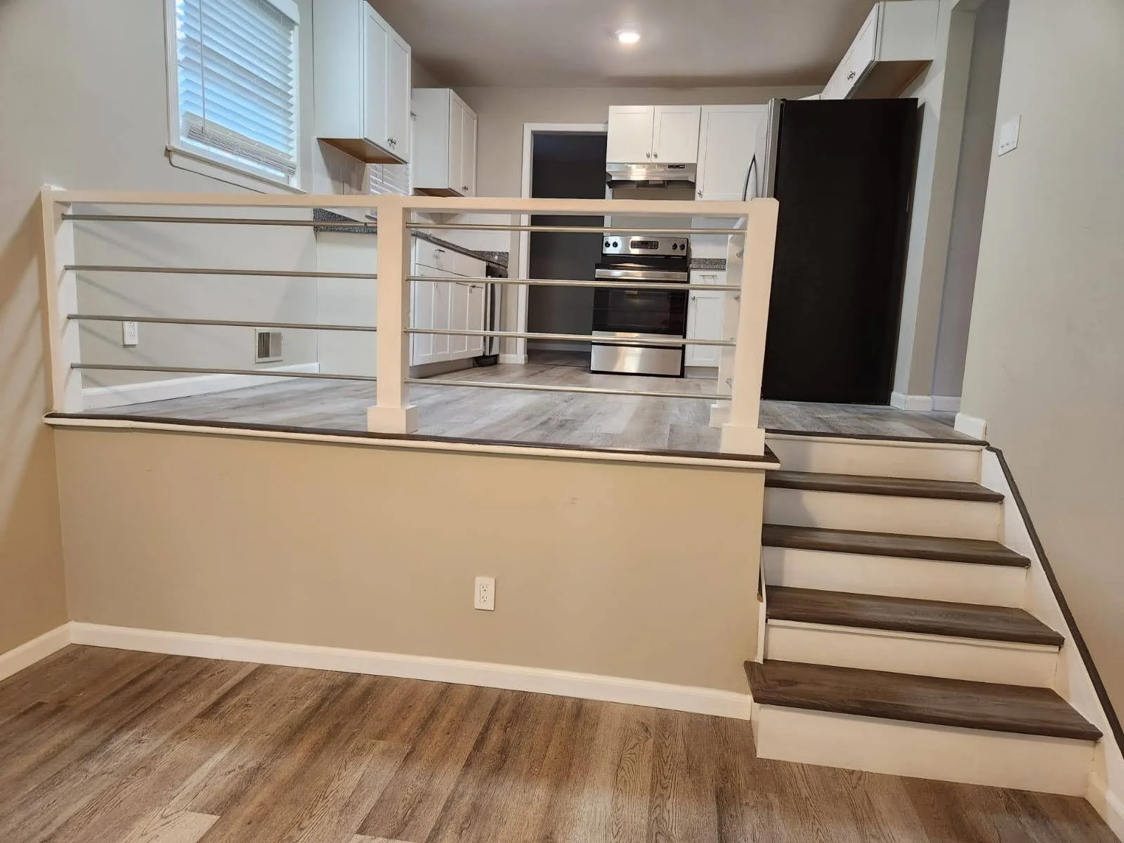 Kitchen with white cabinets, stainless steel oven, black refrigerator, and a small staircase leading to a raised dining area with a railing, beige walls, and wood flooring.