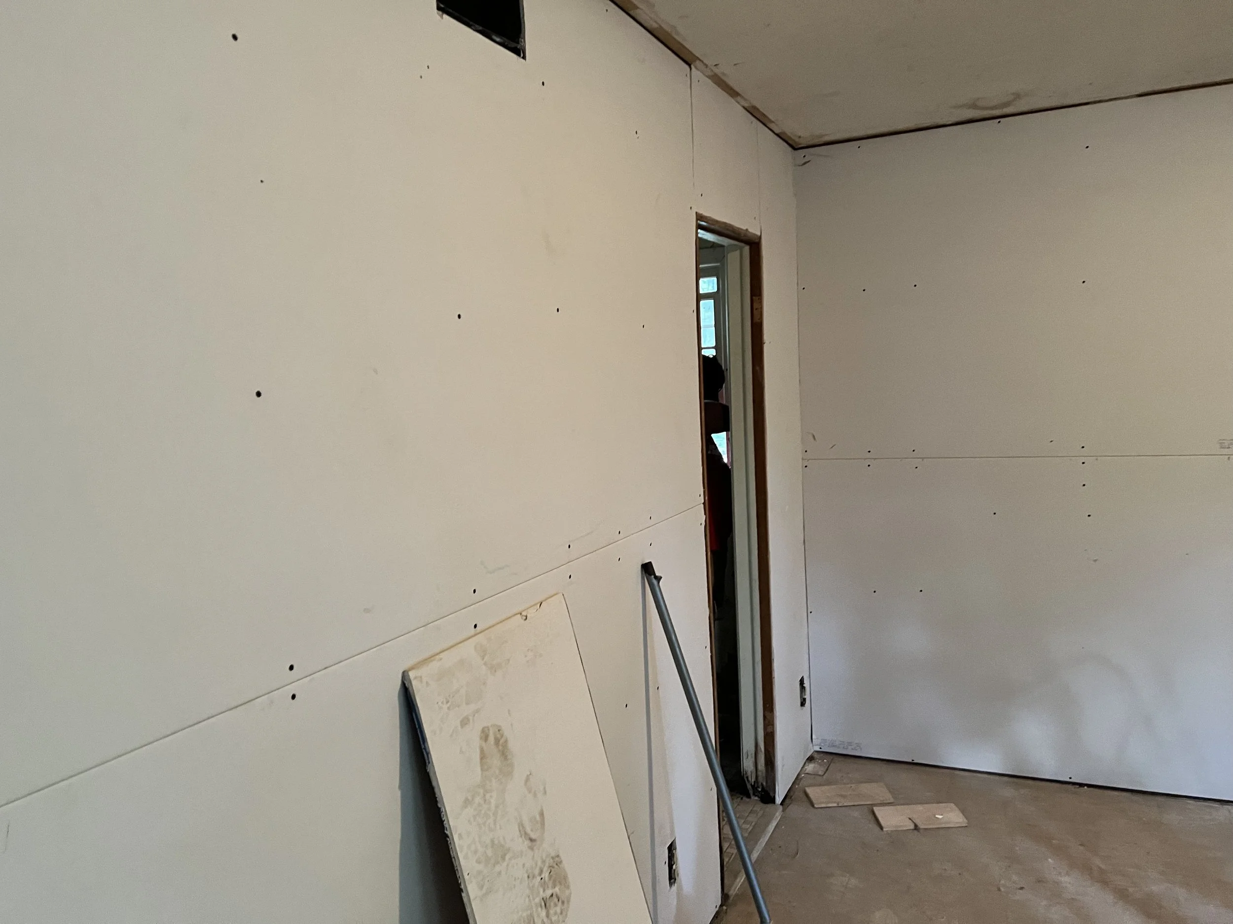 Interior of a room under construction with drywall sheets on the walls, a leaning piece of drywall, a mop handle, and a person visible through a doorway in the background.