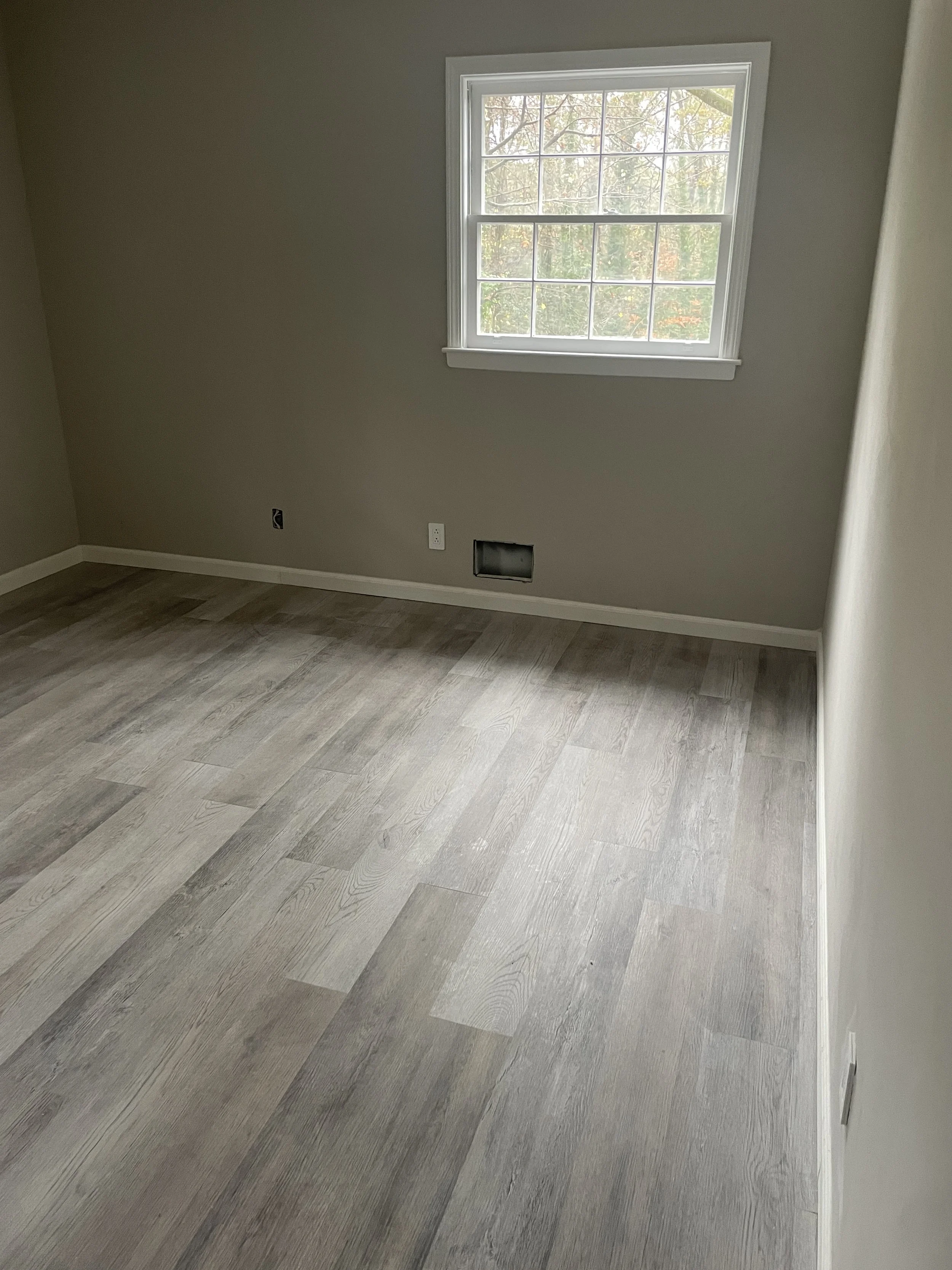 Empty room with beige walls, light wood flooring, a window showing trees outside, and an air vent in the wall.