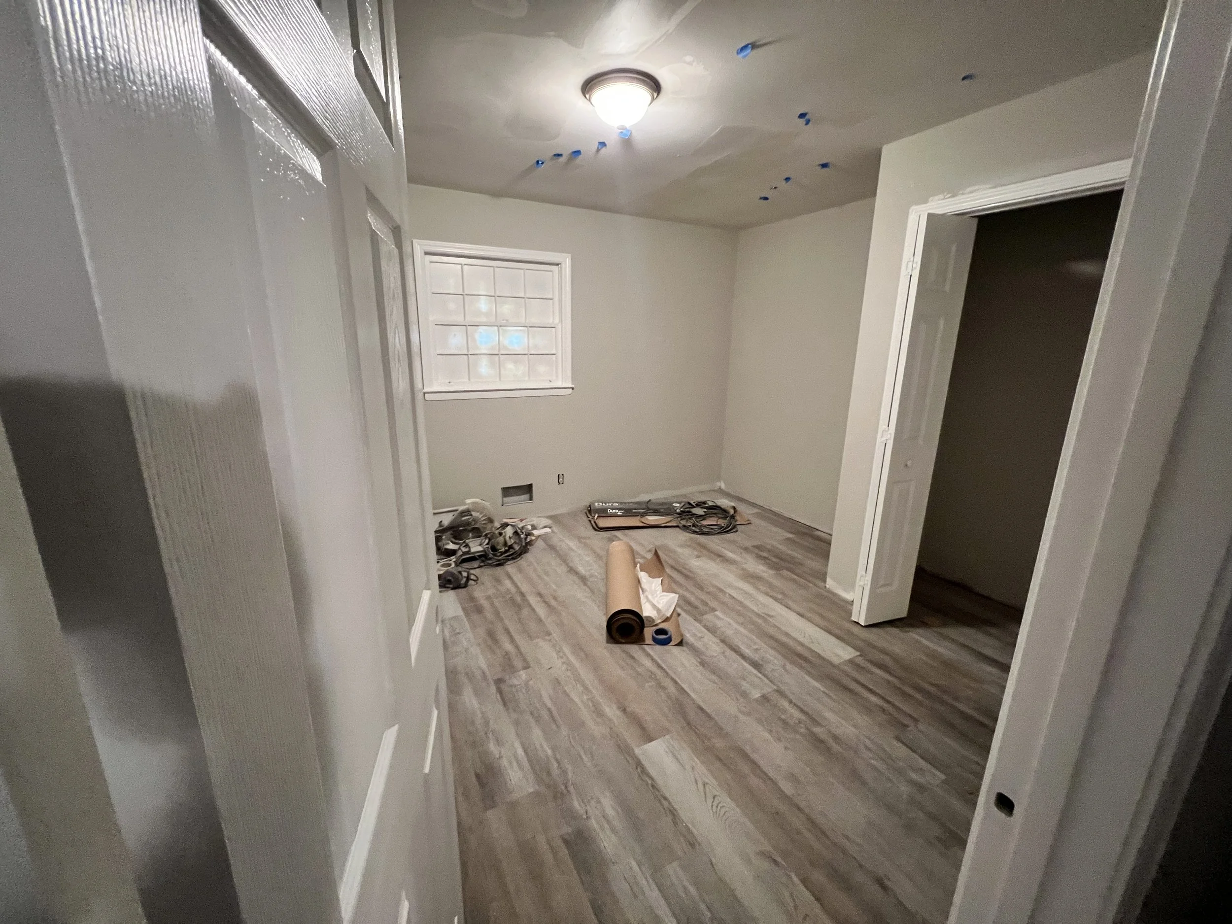 Empty room under renovation with wood flooring, unfinished walls, a small window, and construction tools and materials on the floor.
