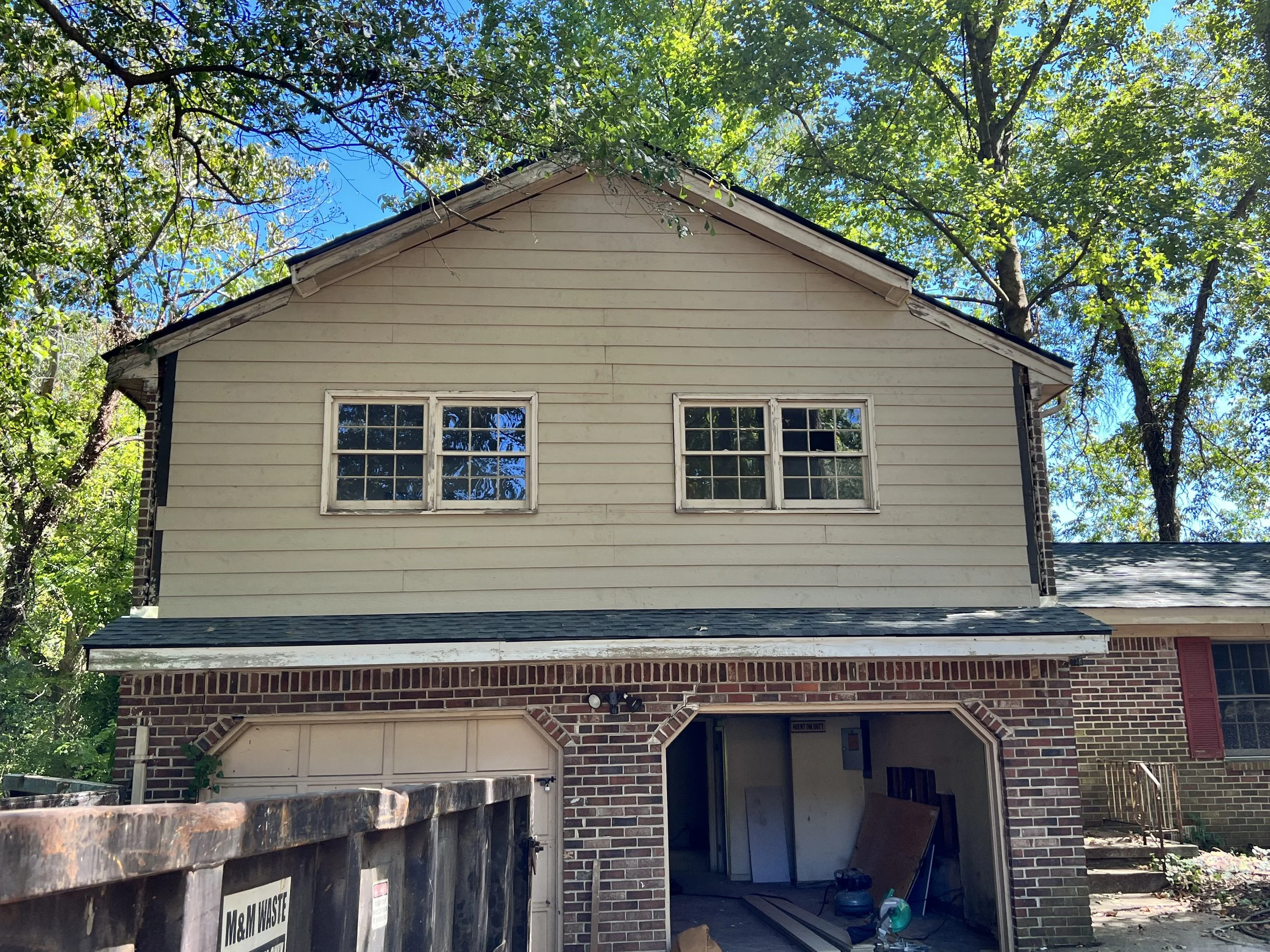A two-story house under renovation, with brick and beige siding exterior, two windows on the upper level, a garage on the left, and construction tools inside the garage opening.
