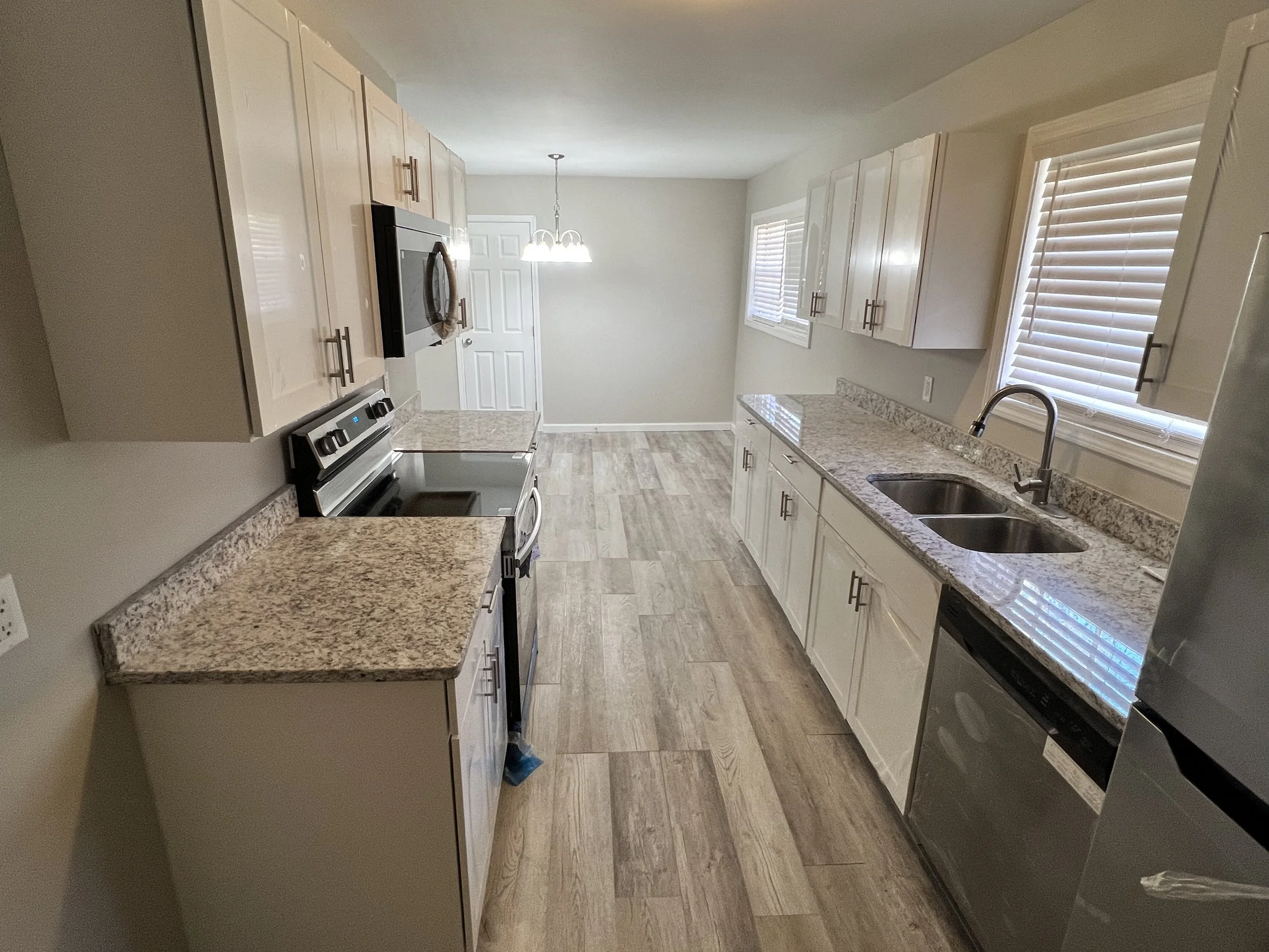Kitchen with granite countertops, white cabinets, stainless steel appliances, including a microwave, dishwasher, and stove. Double sink near a window with blinds, and a dining area with a hanging light fixture at the back.