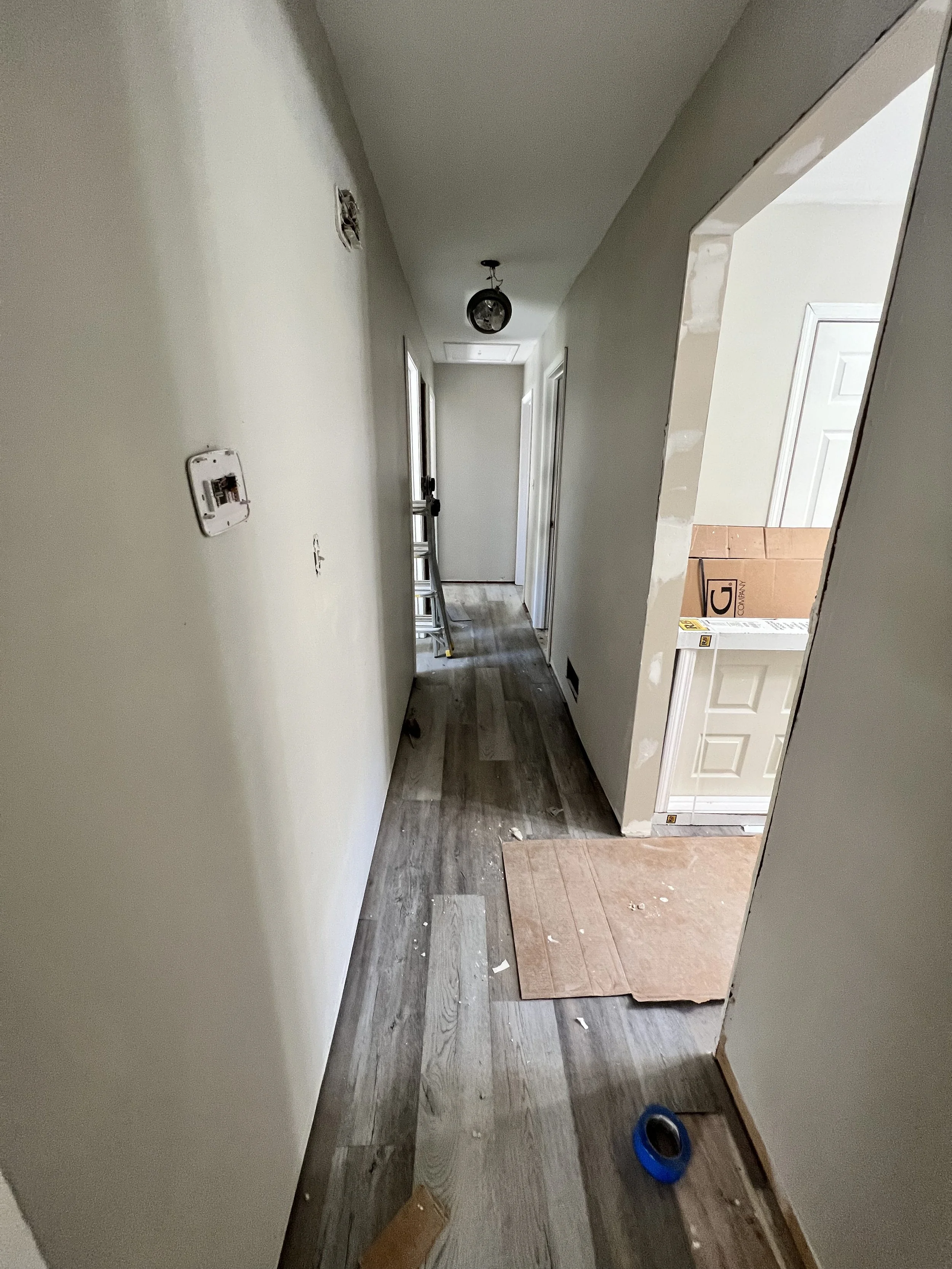 Interior hallway undergoing renovation, with new gray wood flooring, a ladder, and a partially taped wall leading to a door at the end.