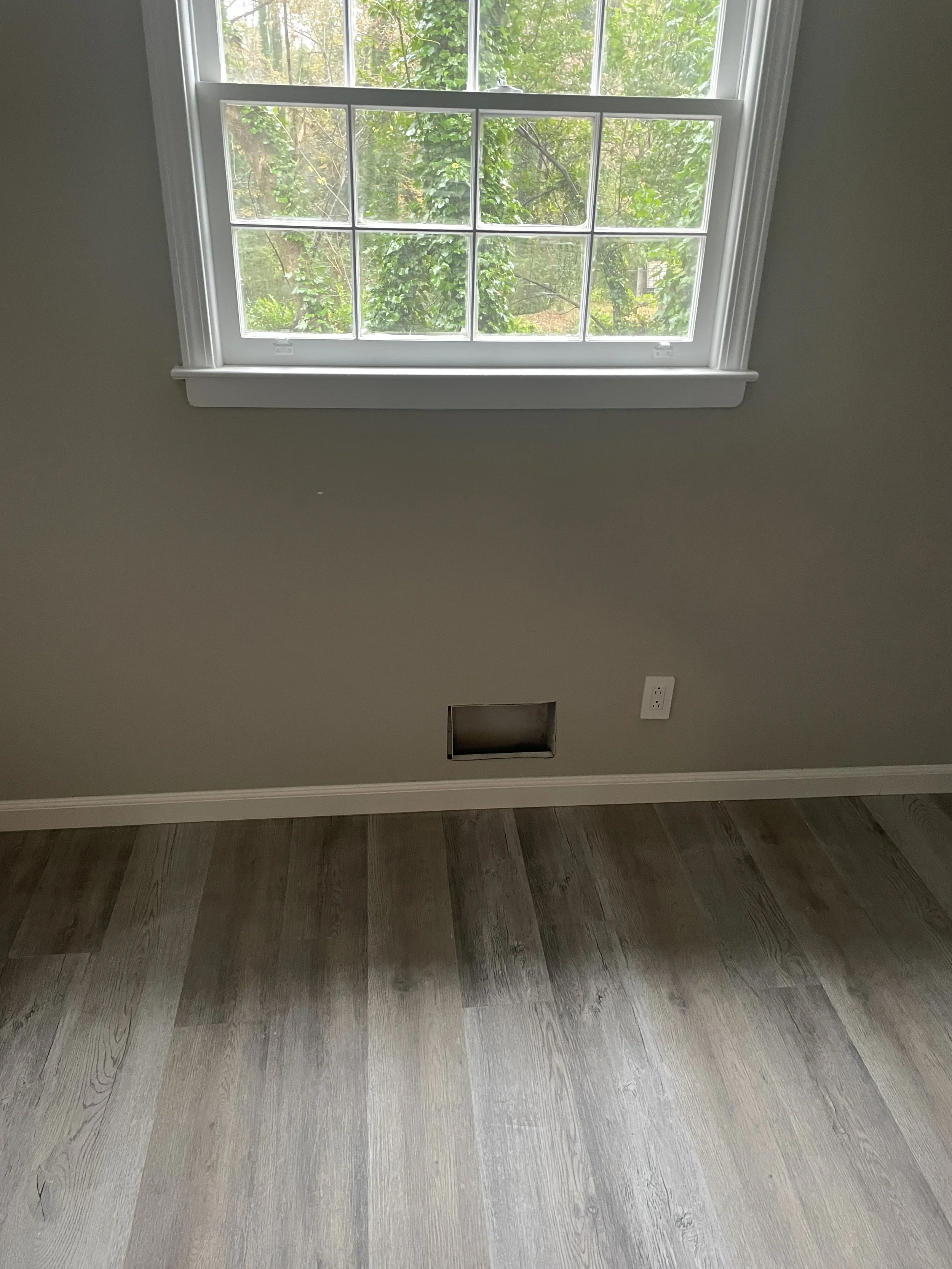 Empty room with a window showing greenery outside, a vent opening in the wall, and an electrical outlet next to it.