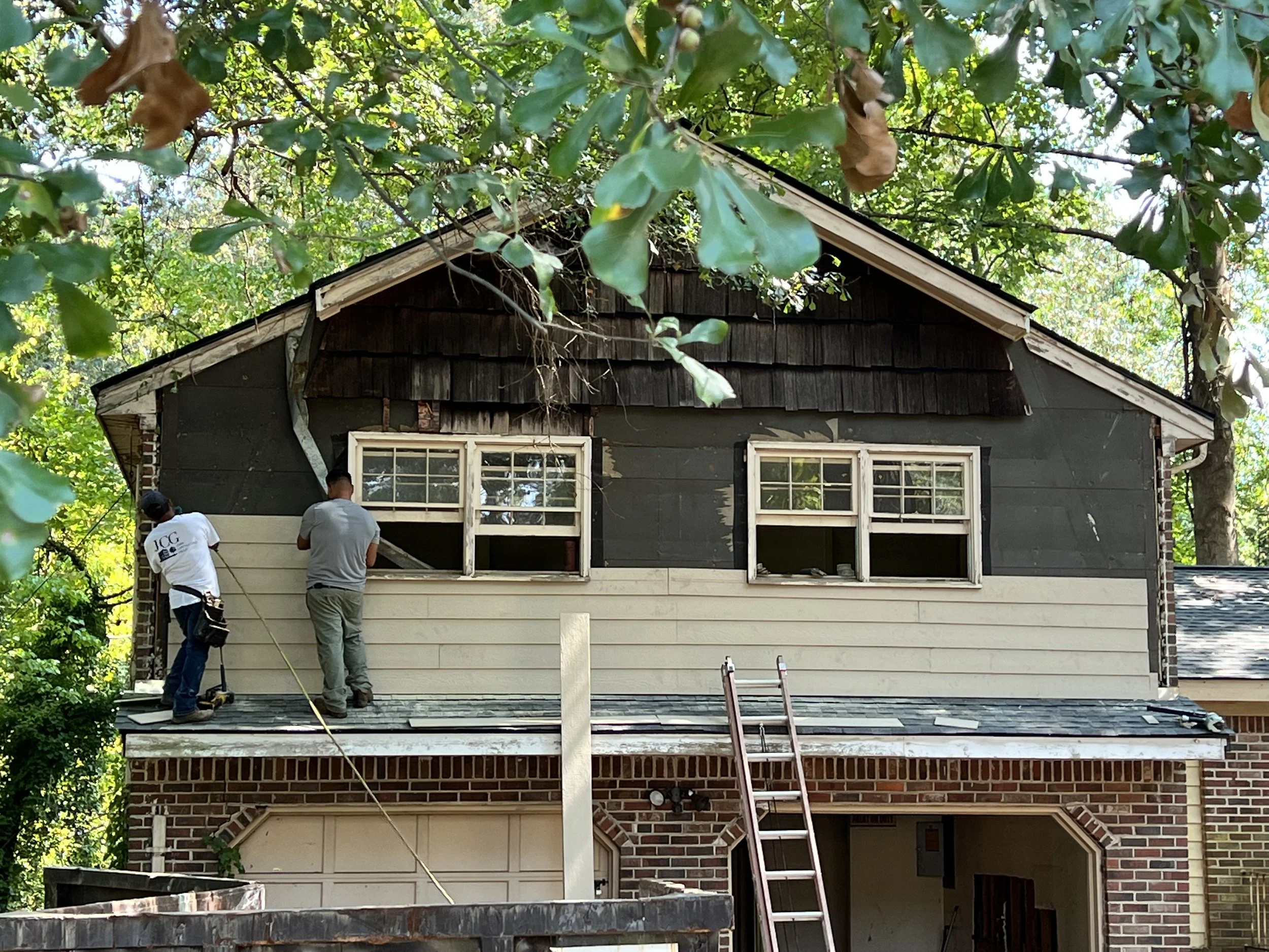 Two workers are repairing the second story of a house with a mix of black and beige siding, and visible windows. The house is surrounded by green trees.