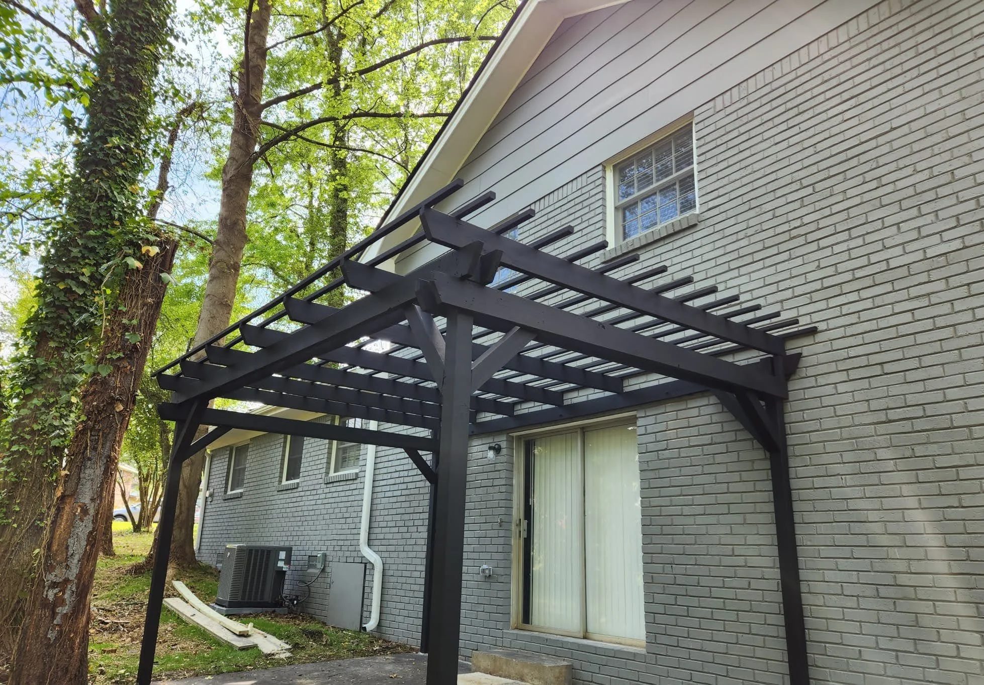 Black wooden pergola attached to a gray brick house with a small window, surrounded by green trees.