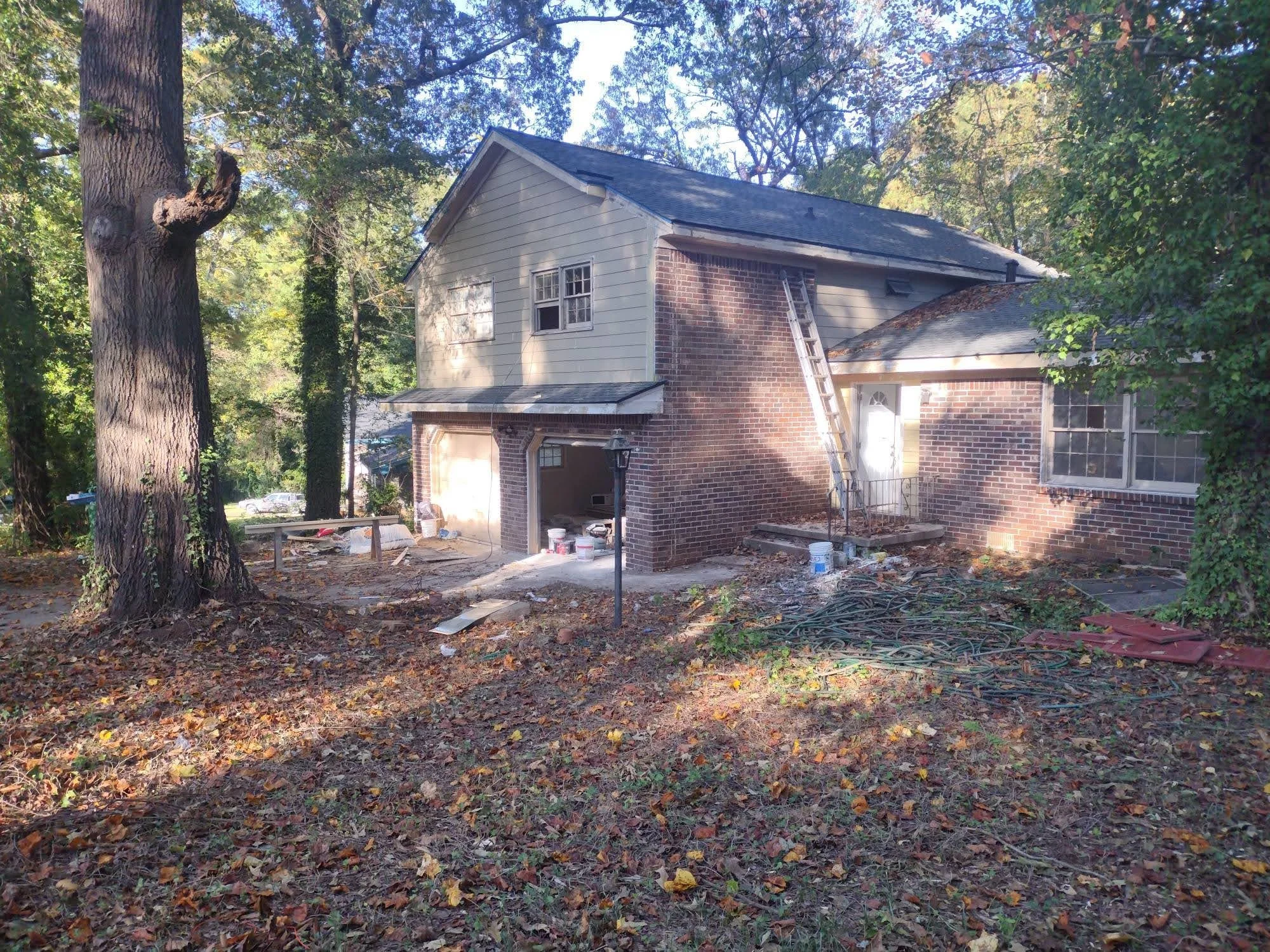 Undergoing construction or renovation, the backyard of a house with a ladder against the wall, construction materials, and scattered debris on the ground, surrounded by trees.