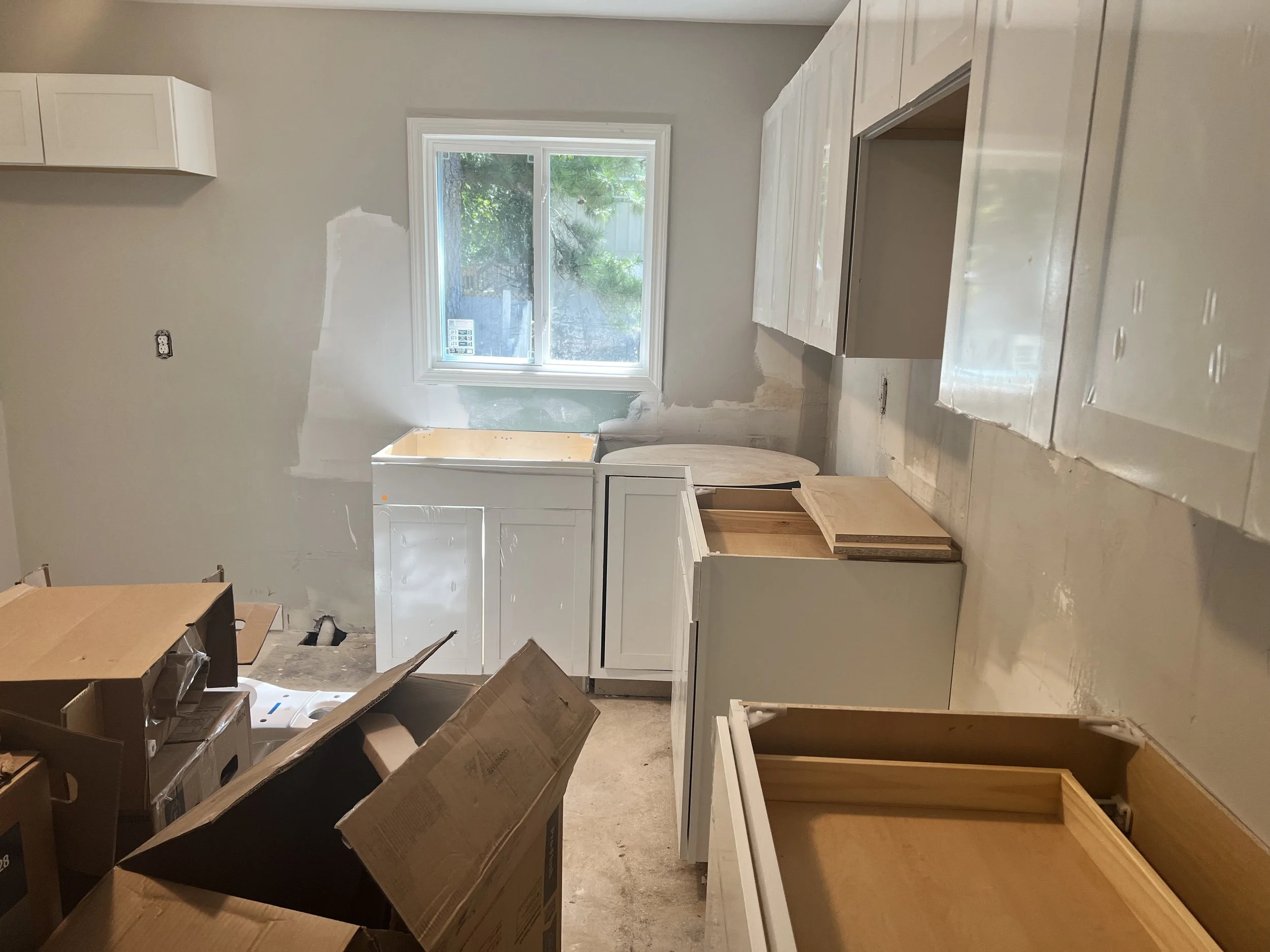 Kitchen under construction with unfinished cabinets, boxes, and a window with trees outside.