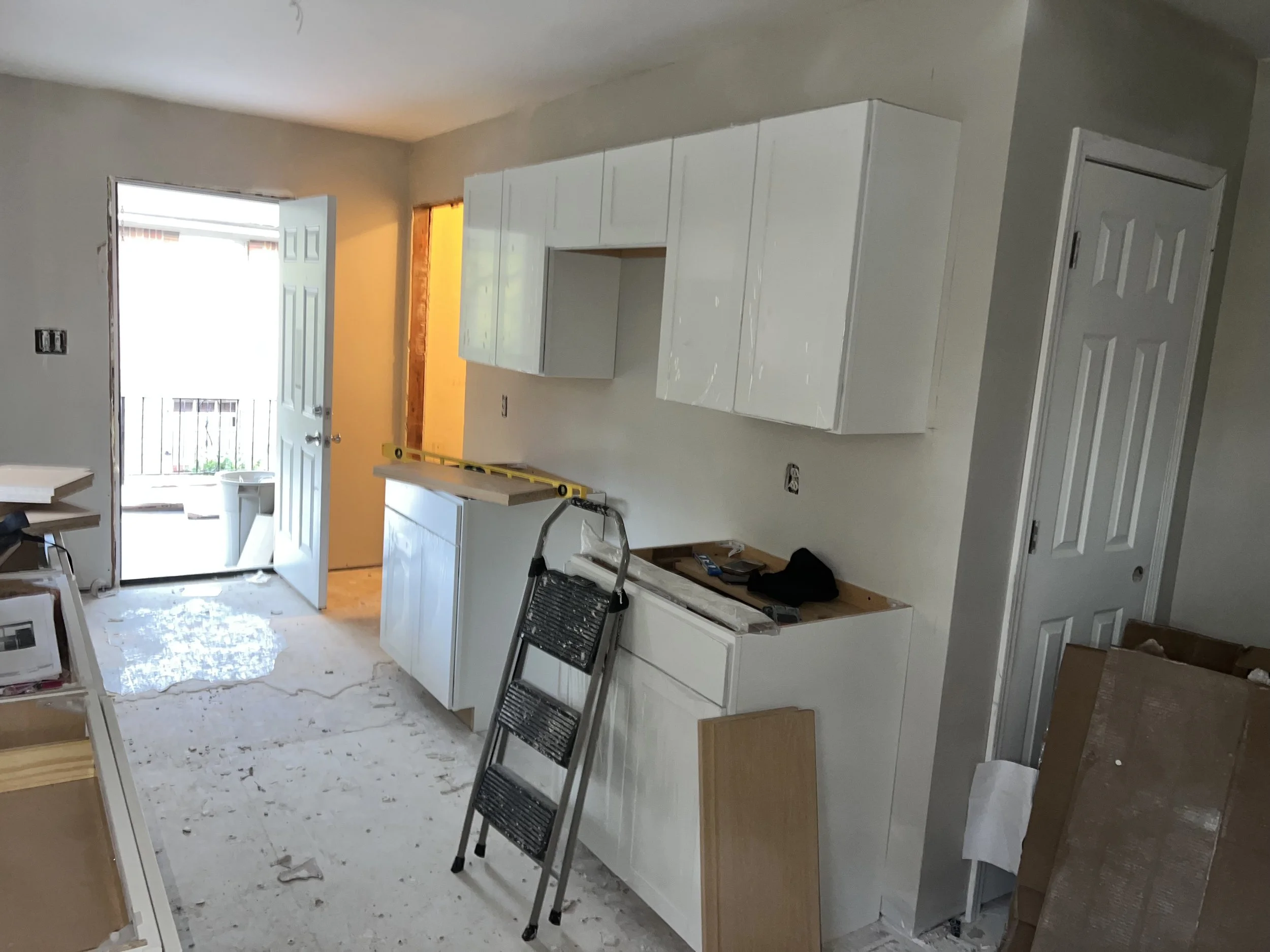 Kitchen under construction with white cabinets, a ladder, and construction tools, leading to a door with a balcony and a small enclosed outdoor area.