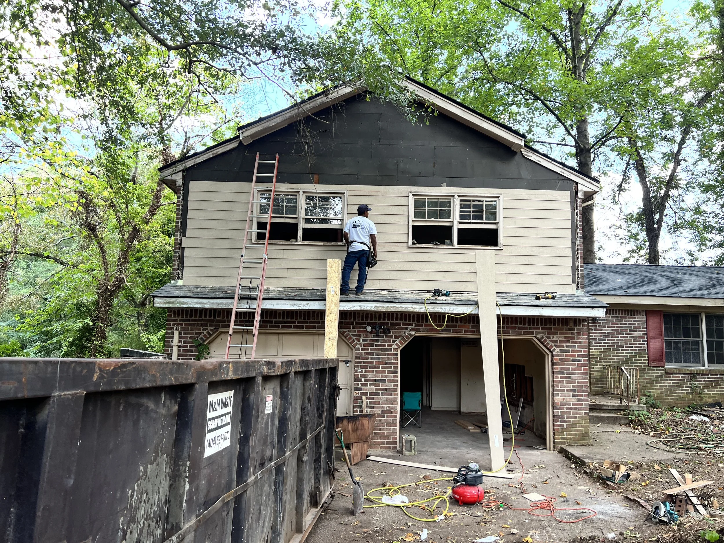 Home renovation in progress with a worker on the second floor installing new siding, ladders, construction tools, and debris in the driveway.