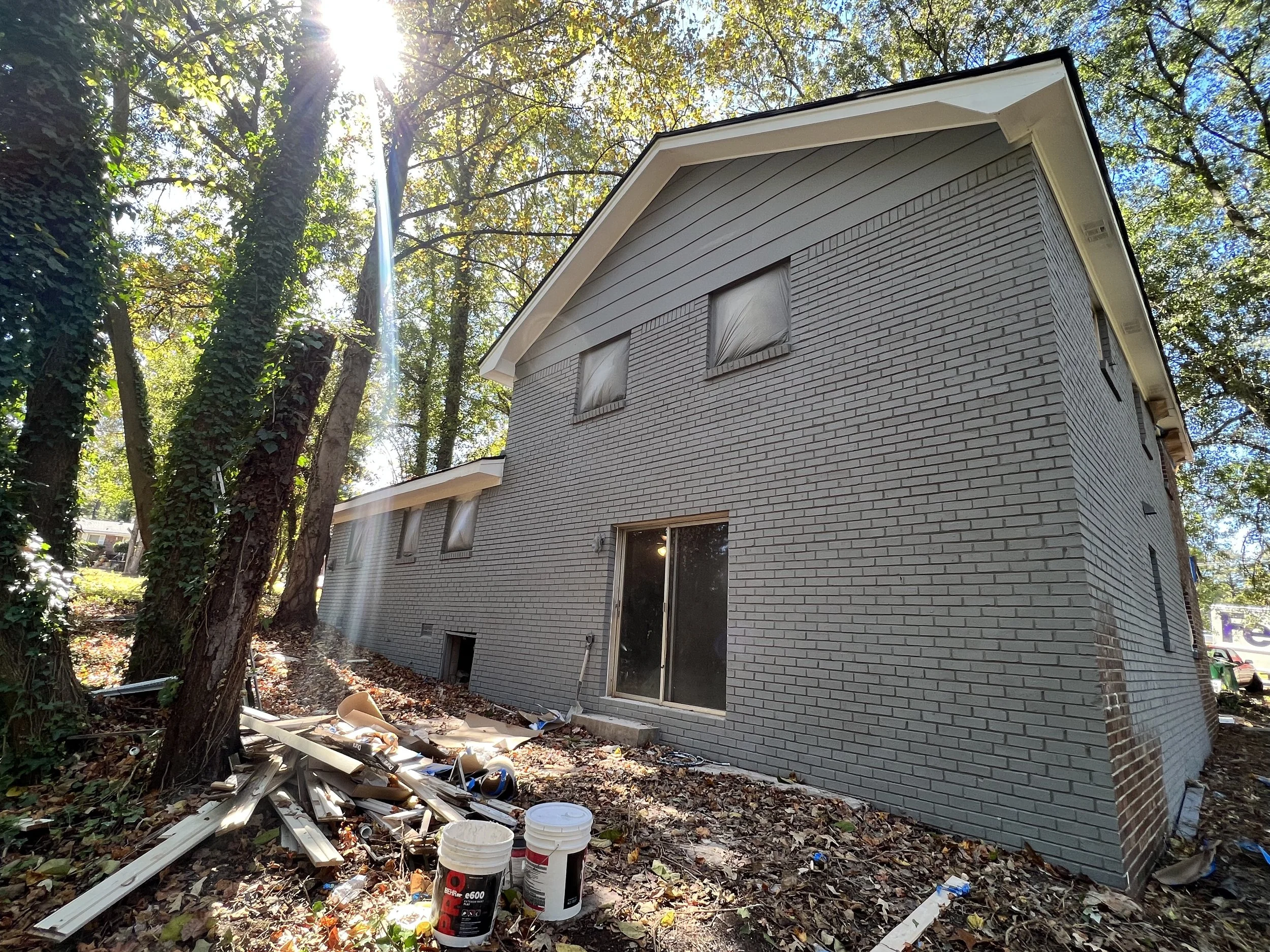 A house under construction with a gray brick exterior, surrounded by trees, and construction debris on the ground.