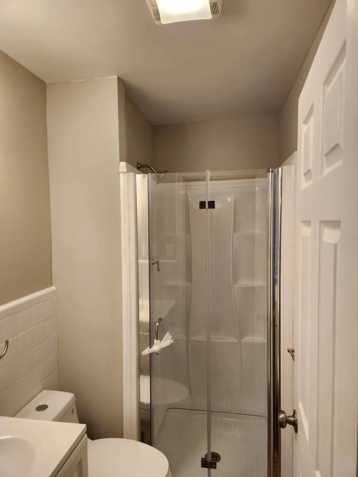 Small bathroom with a walk-in shower with a clear glass door, a toilet, a beige tiled wall, and a closed white door.