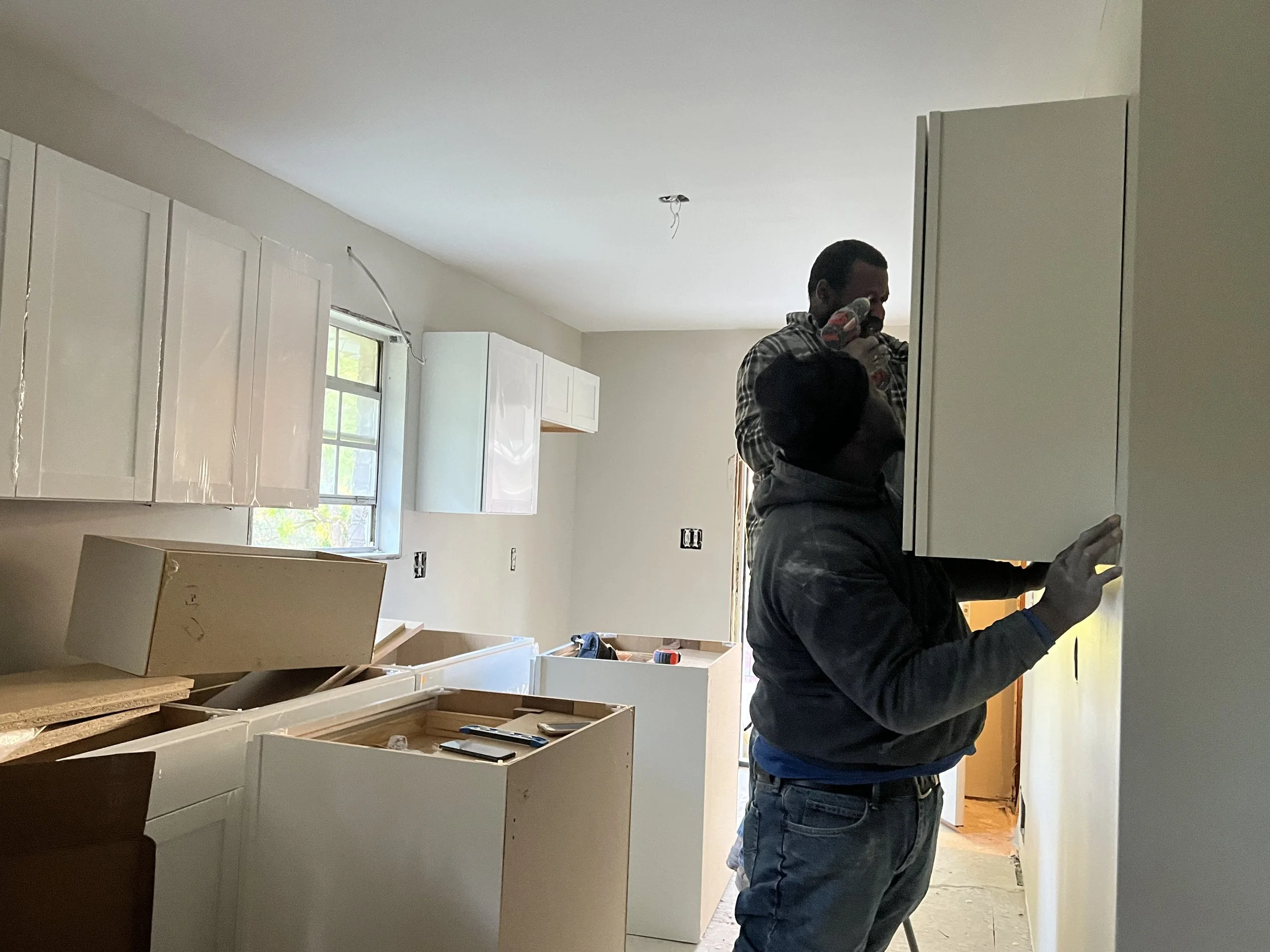 Two men are working on a home renovation or construction project. One man is using a power drill to install or work on kitchen cabinets. The room is in the process of being finished, with some construction materials, cabinets, and tools visible.