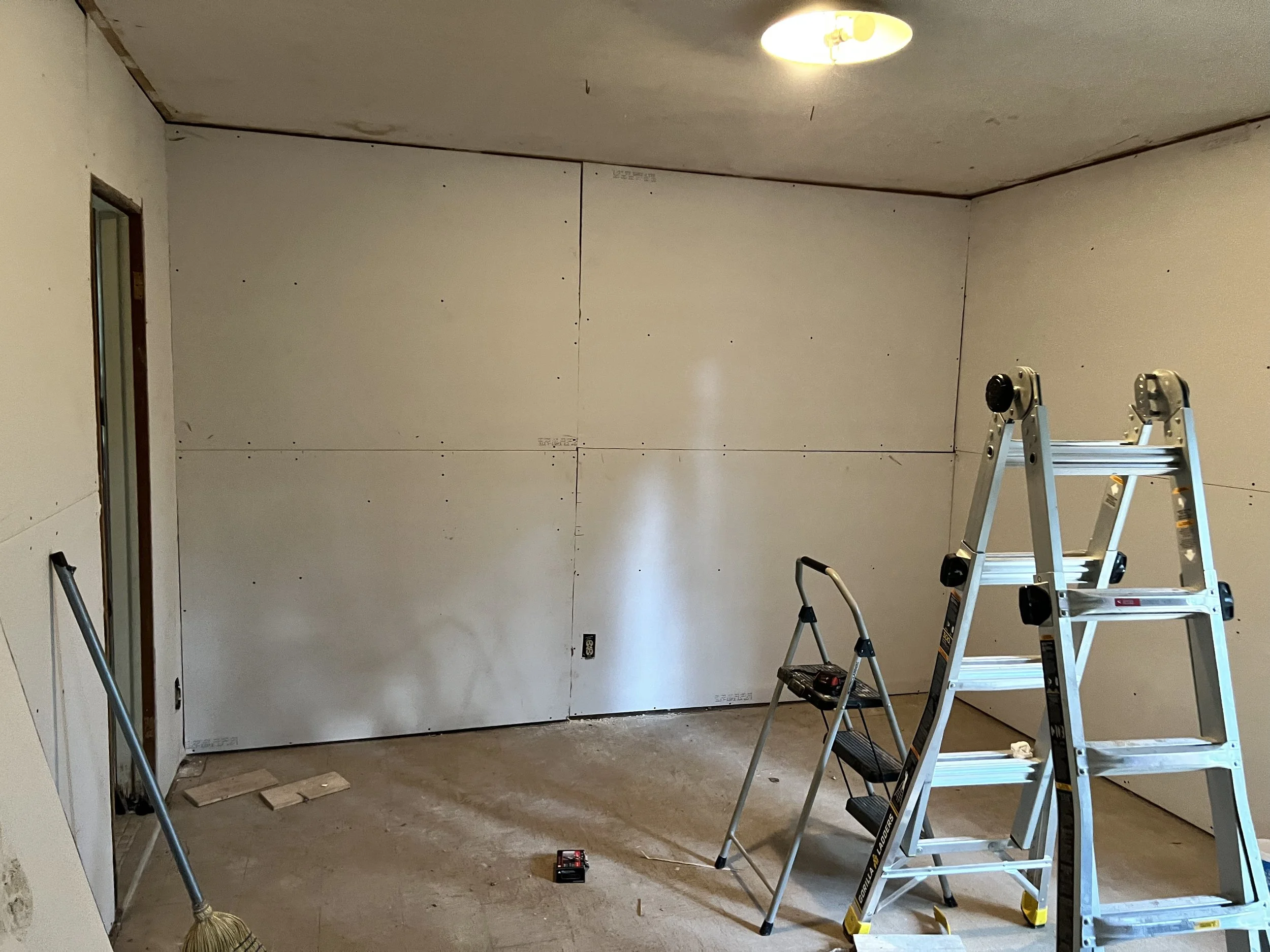 Room under renovation with unfinished drywall, a ladder, a step stool, a small toolbox, and construction tools along with a ceiling light fixture.