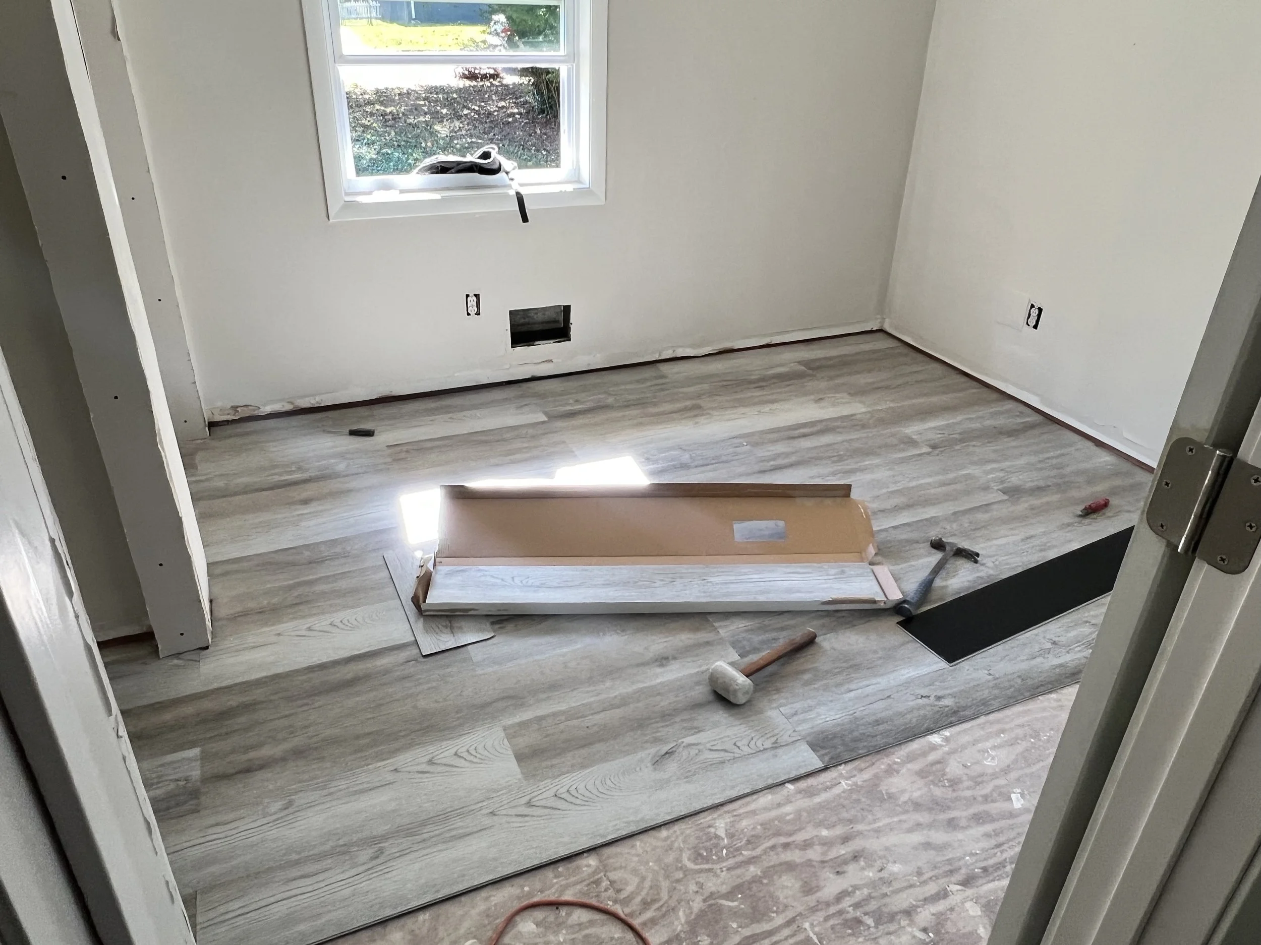Room under construction with newly installed wood laminate flooring, tools like hammer and pry bar on the floor, window with a view of outside, electrical outlets and a vent on the wall.