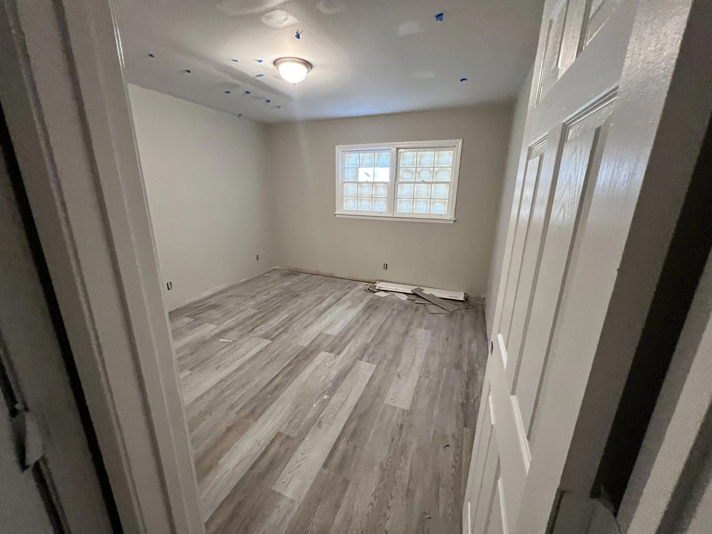 Unfinished empty room with a window, hardwood floors, and a ceiling light fixture. Construction materials are on the floor near the wall.