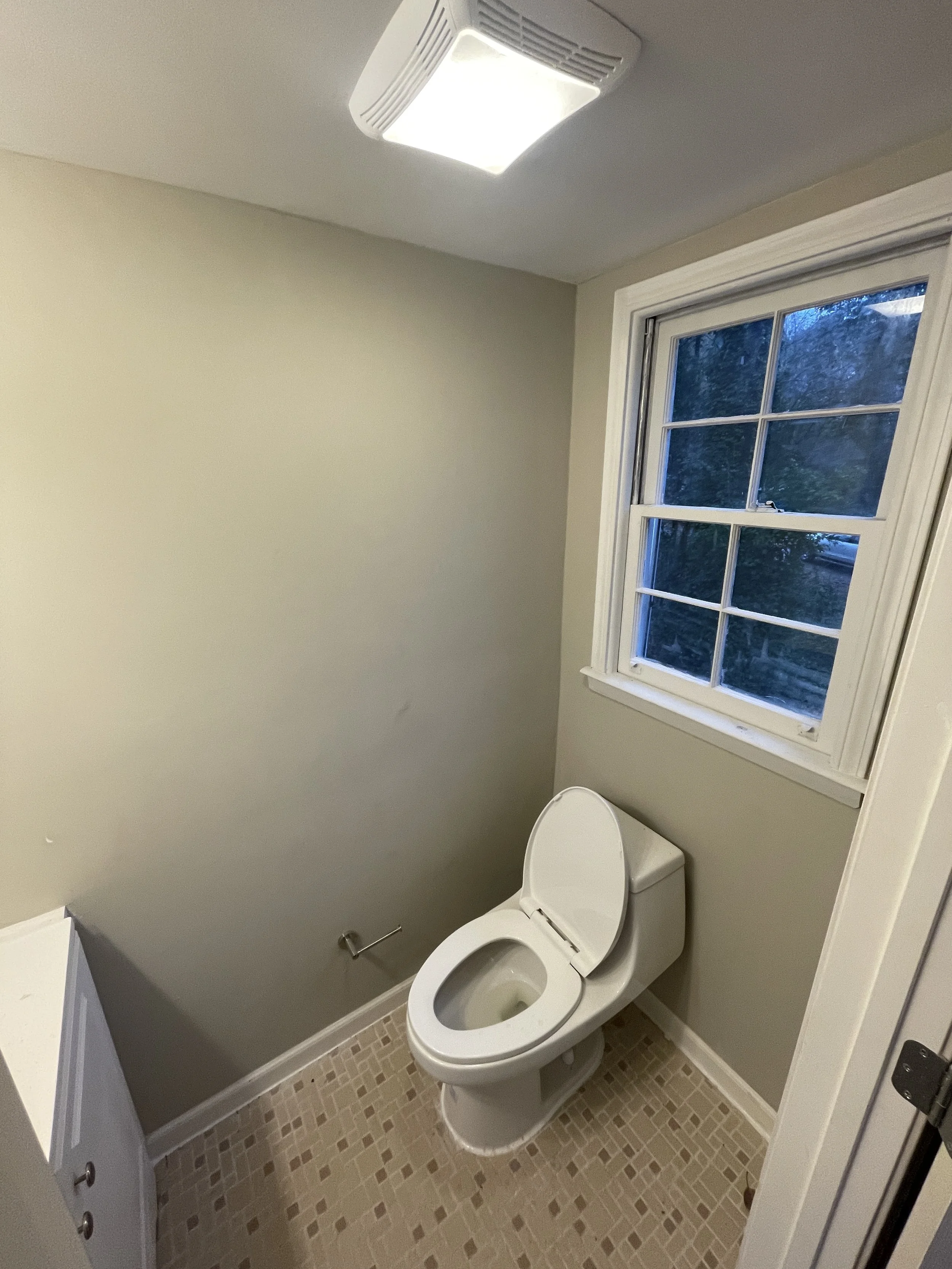 A small bathroom with a white toilet, a window above it, beige tiles on the floor, and a ceiling light.
