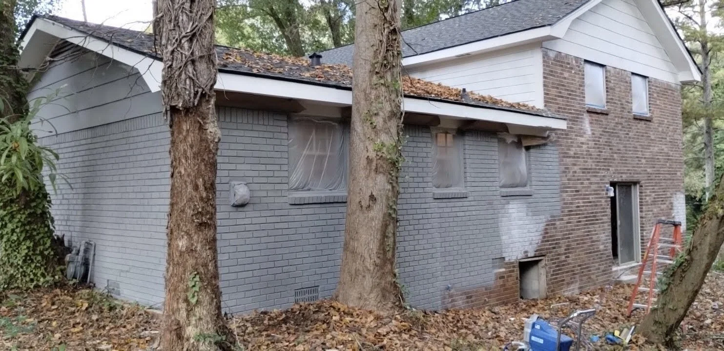 A house under renovation, with its lower brick exterior partially painted in gray, surrounded by trees and construction tools on the ground.