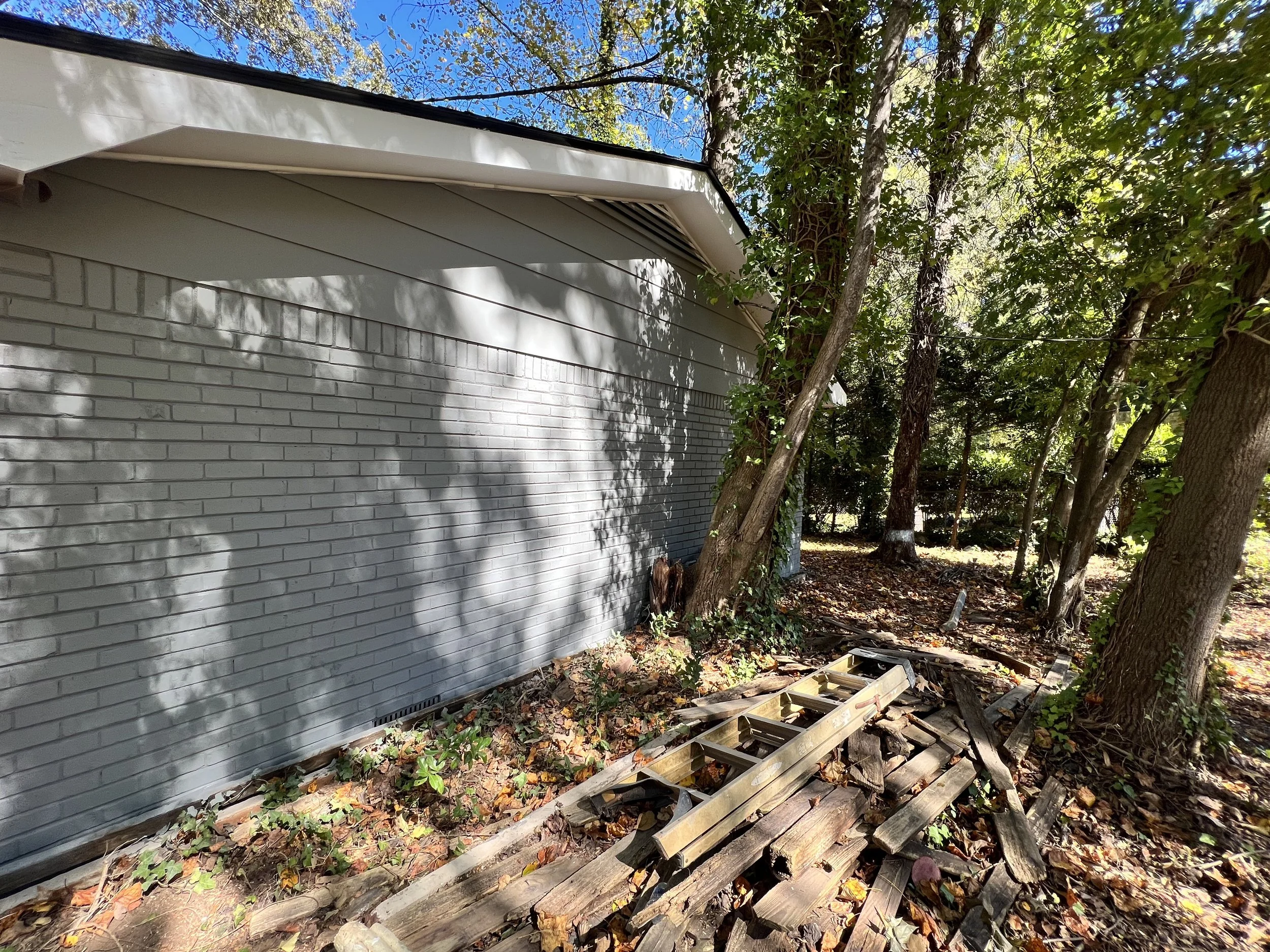 Side of a house with white brick wall, surrounded by trees with green leaves, in a wooded yard. Construction debris and wood planks are on the ground.