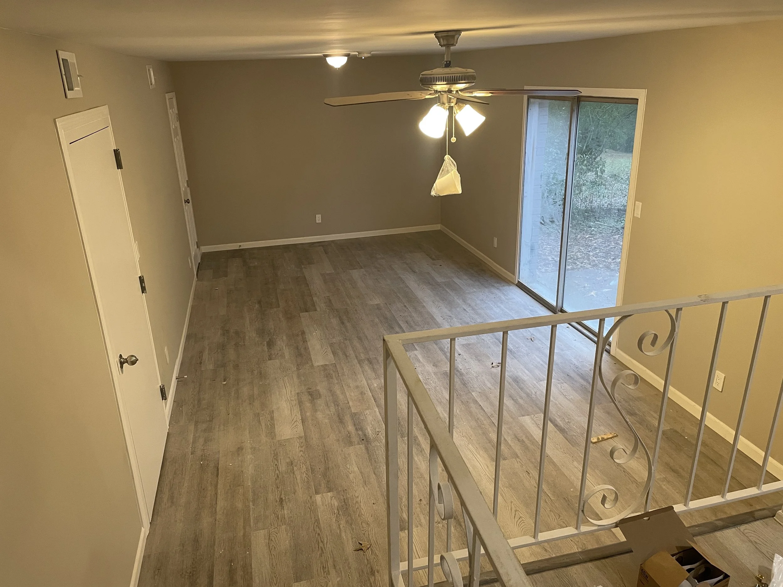 Empty living room with wooden flooring, a ceiling fan, a sliding glass door leading outside, and a staircase with a decorative white railing.