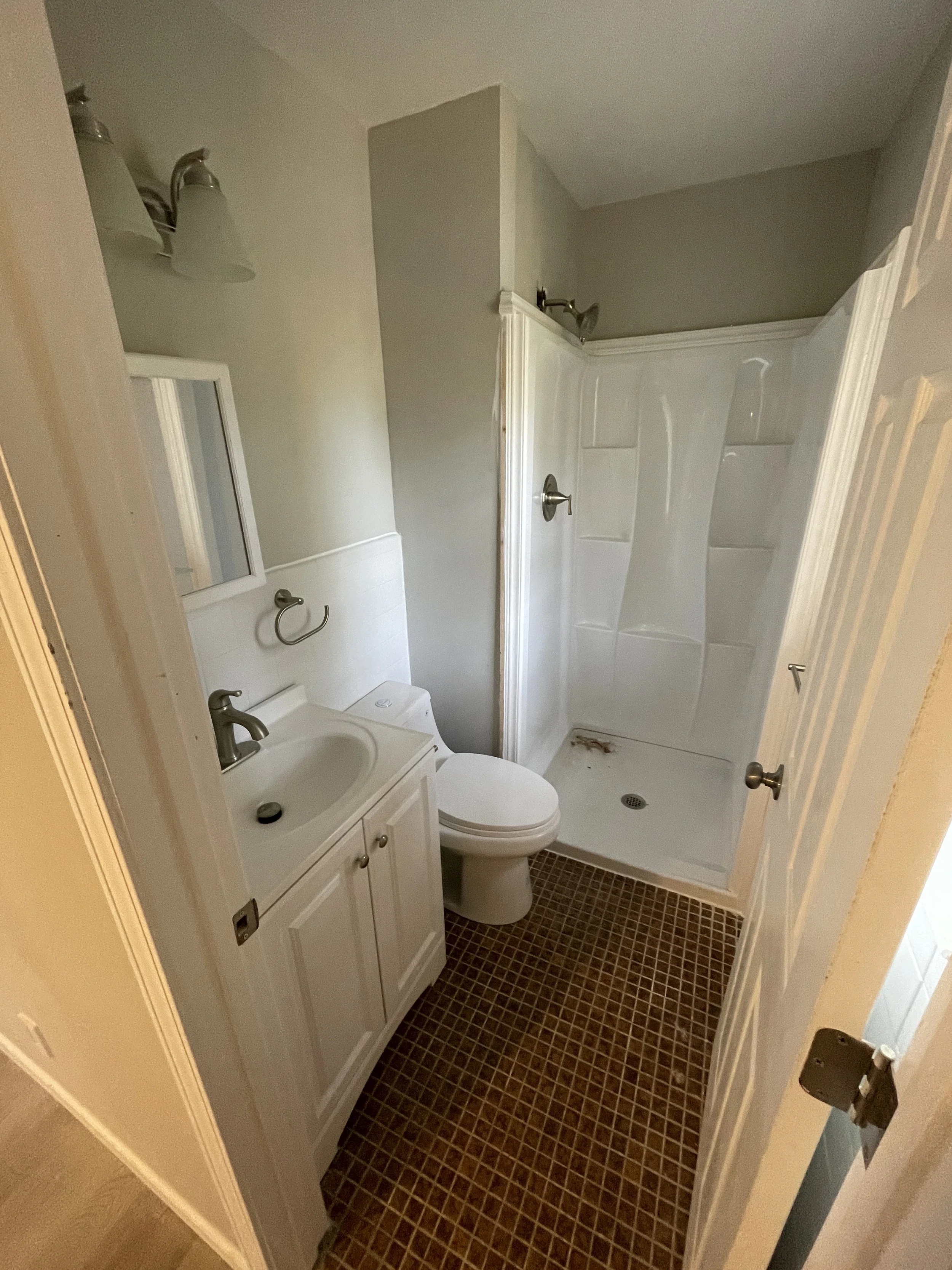 Small bathroom with a white vanity sink, toilet, and shower stall with glass door. The floor is covered in small brown tiles, and the walls are painted light gray.
