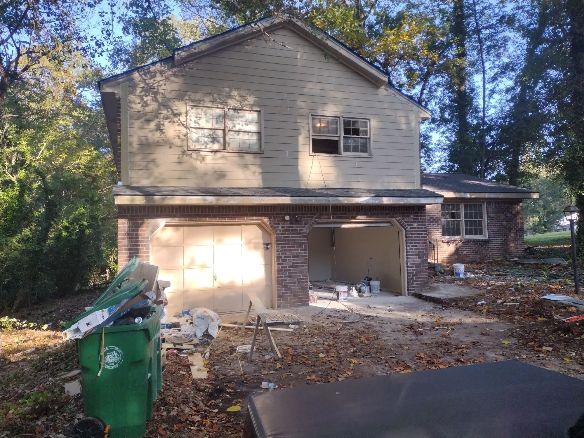 Undergoing house renovation, two-story house with brick and siding exterior, construction tools, buckets, and debris in front, surrounded by trees, driveway leading to garage