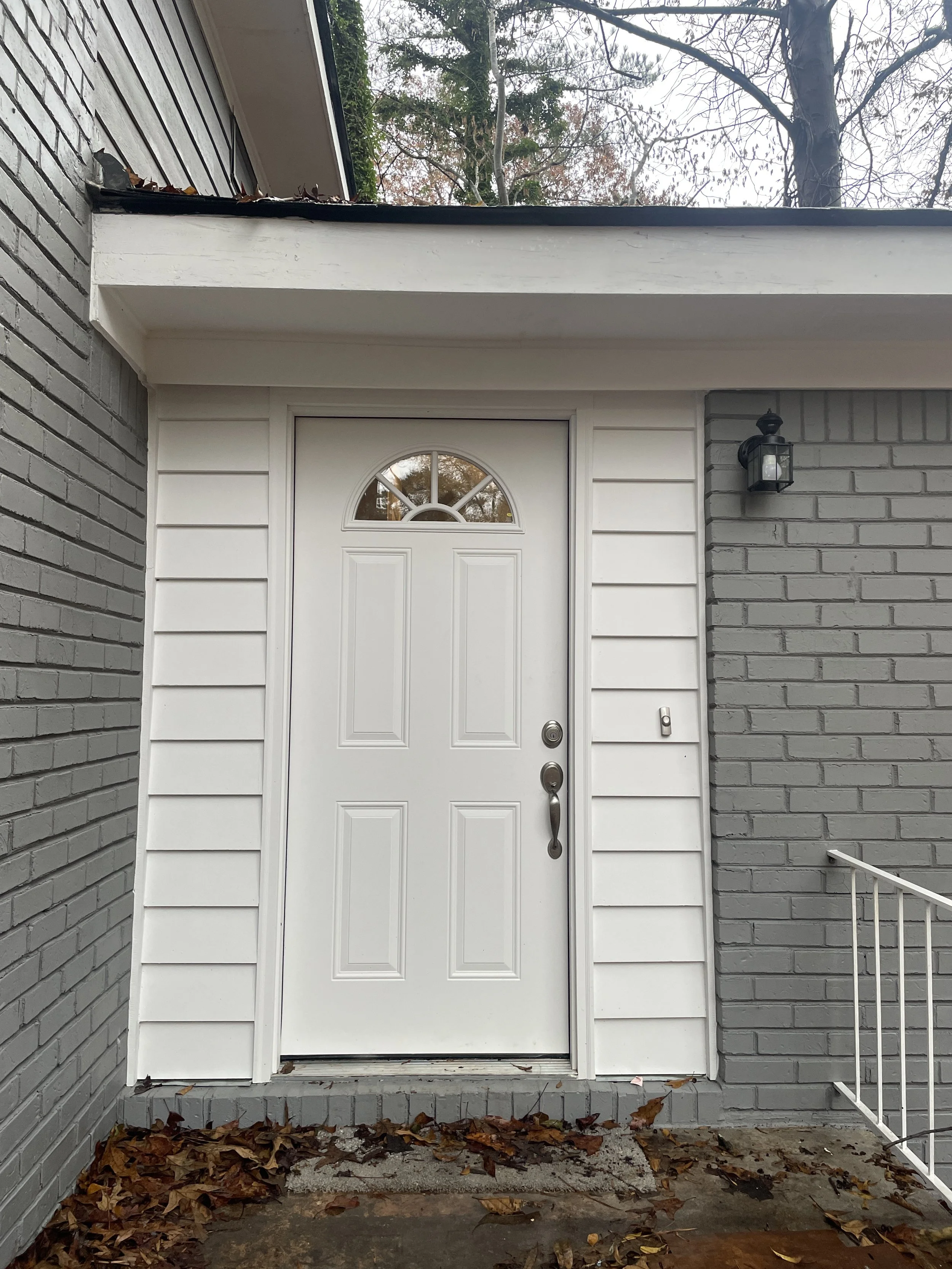 White front door with a small arched window at the top, set in a house with grey brick and white siding, with a black lantern-style light fixture on the wall, and fallen leaves on the ground.
